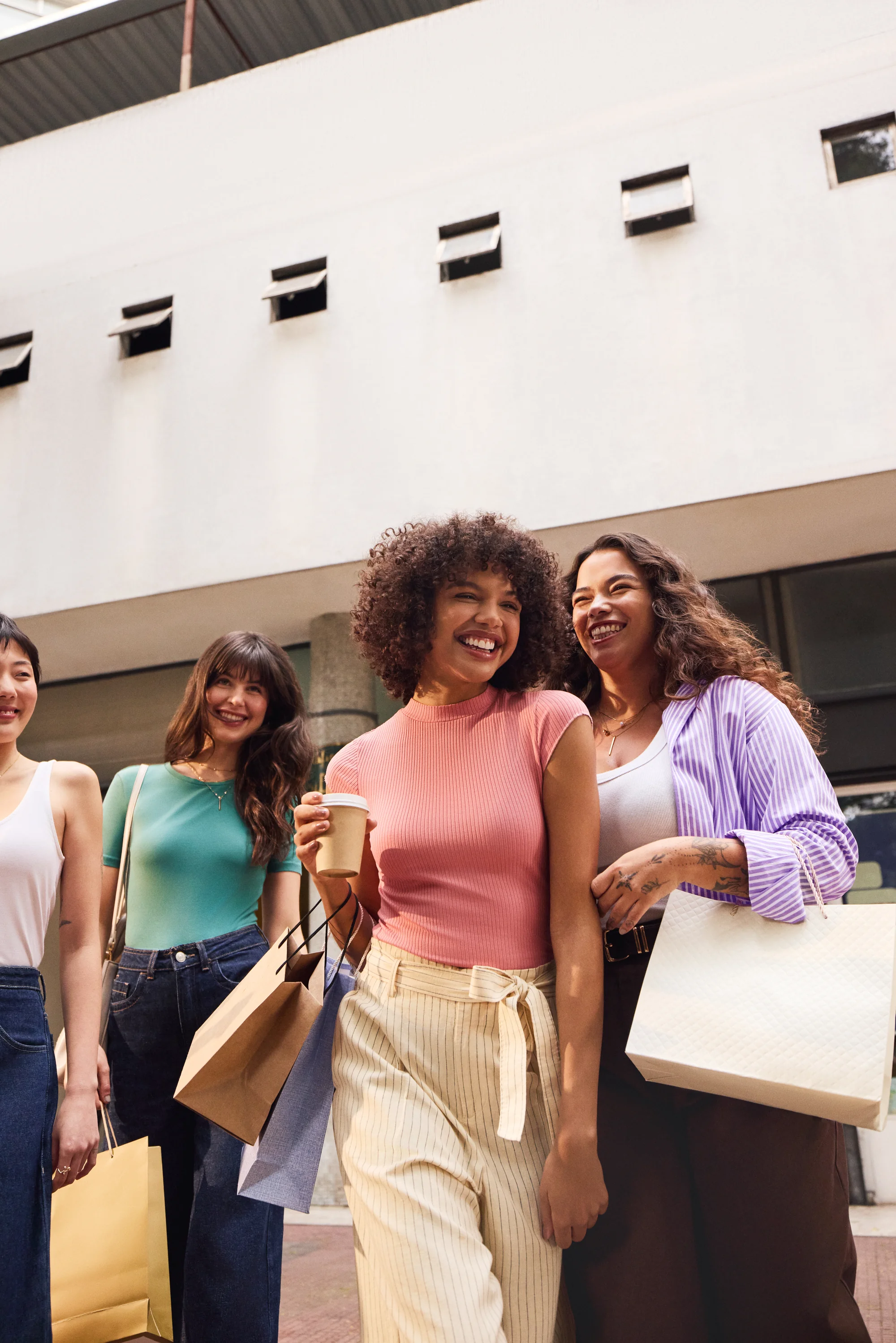 Grupo de amigas sorrindo com sacolas de compras e café.
