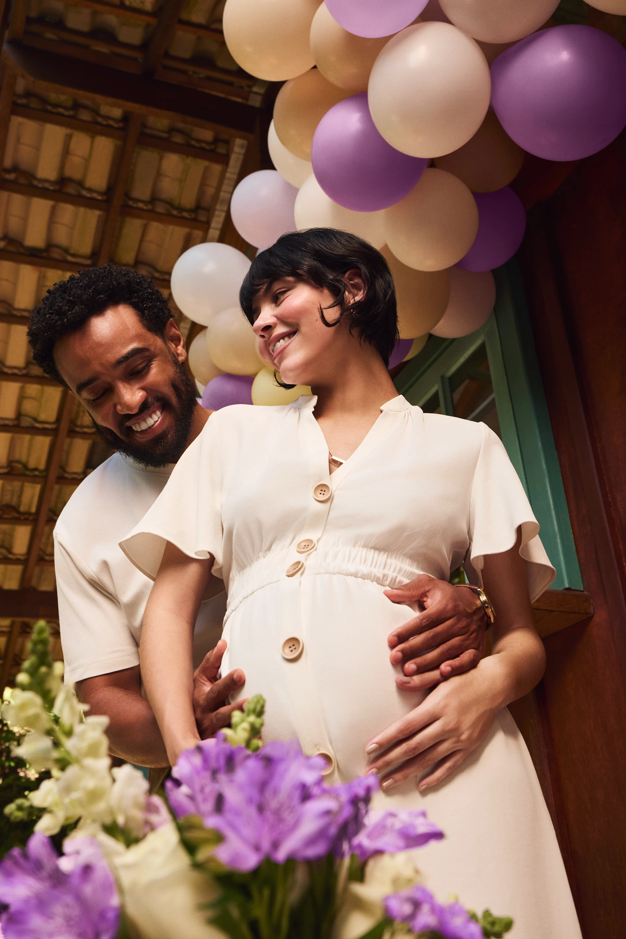 Casal sorridente em ambiente festivo com flores e balões ao fundo.