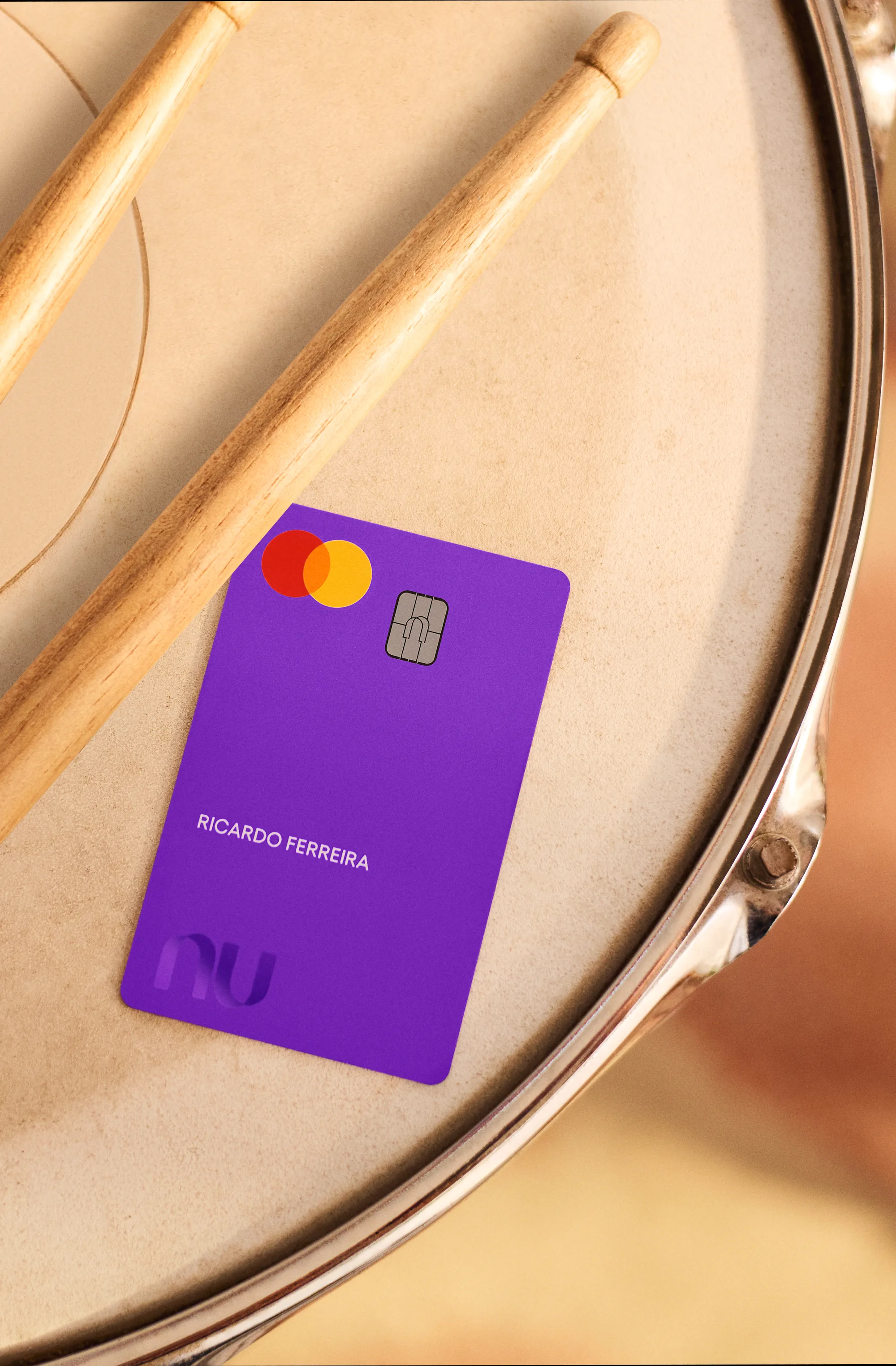 Cartão Nubank roxo em cima de um tambor com baquetas.