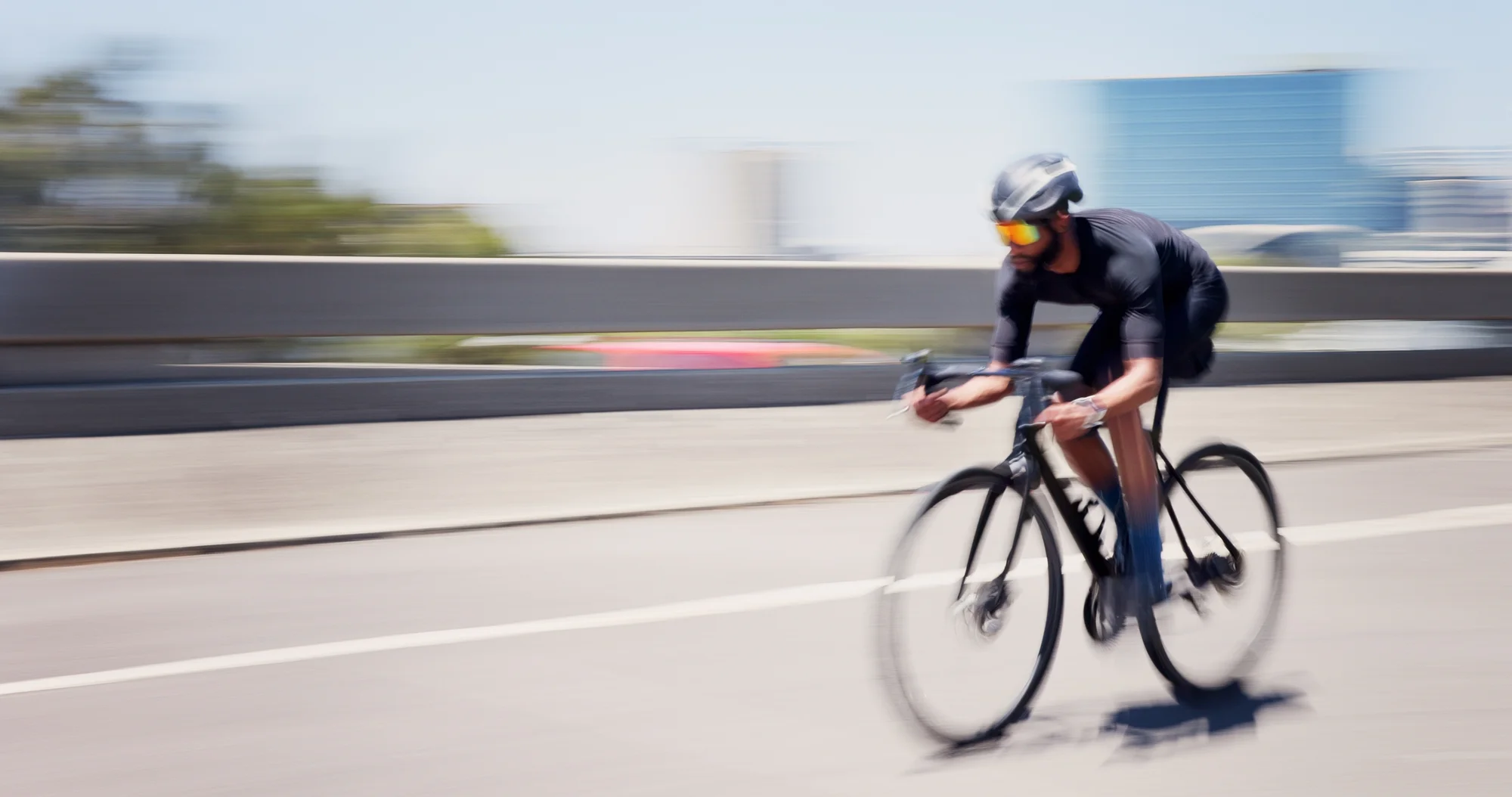 Um ciclista usando uniforme preto, capacete e óculos espelhados coloridos pedala em uma bicicleta de corrida por uma estrada urbana.
