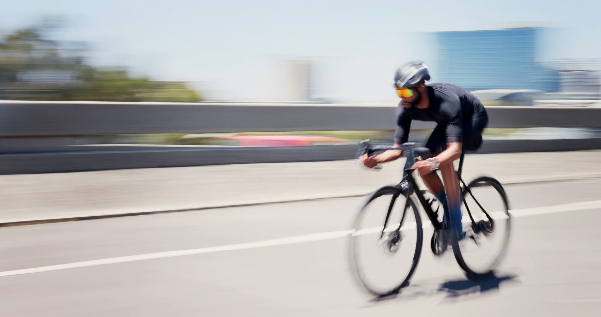 Um ciclista usando uniforme preto, capacete e óculos espelhados coloridos pedala em uma bicicleta de corrida por uma estrada urbana.
