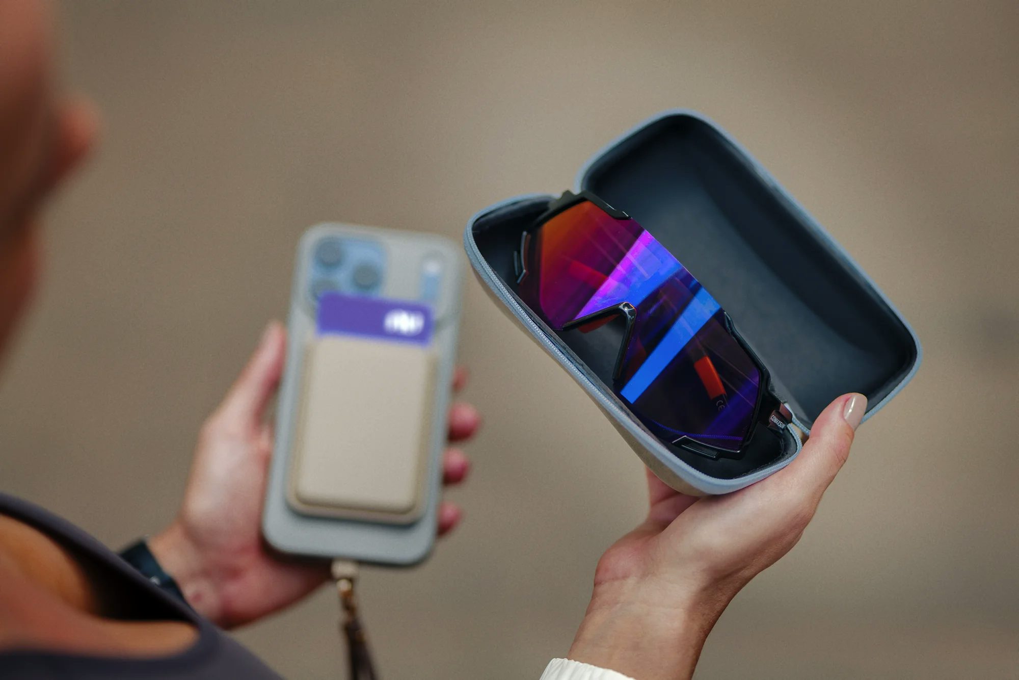Foco nas mãos de uma atleta segurando um estojo aberto com óculos esportivos de lentes espelhadas coloridas. Ao fundo, a outra mão segura um smartphone cinza com um porta-cartões acoplado contendo o cartão Nubank Ultravioleta.