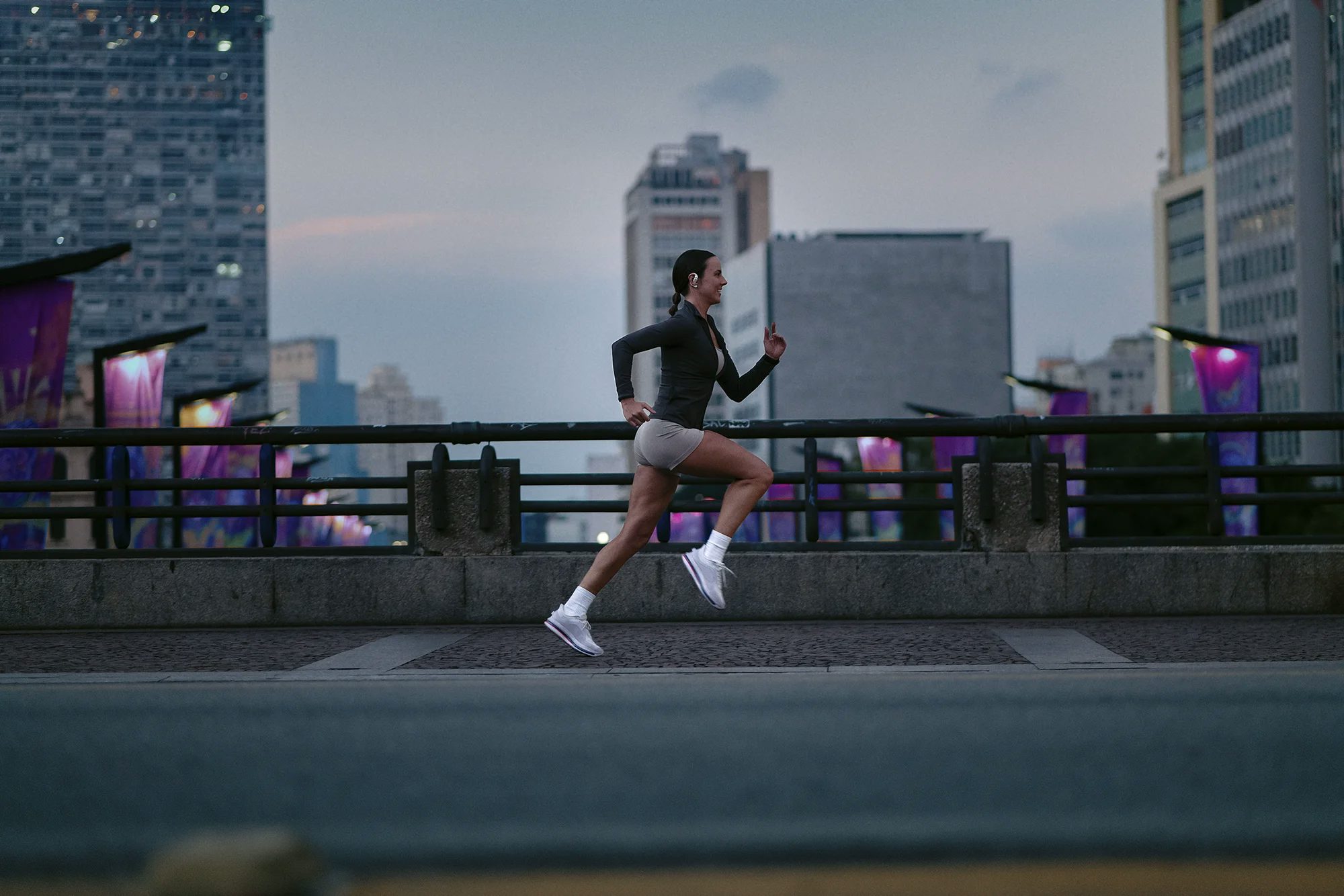 Uma mulher corre ao ar livre sobre uma ponte ou passarela urbana ao entardecer. Ao fundo, observa-se a silhueta de prédios e estruturas com iluminação roxa na paisagem da cidade.