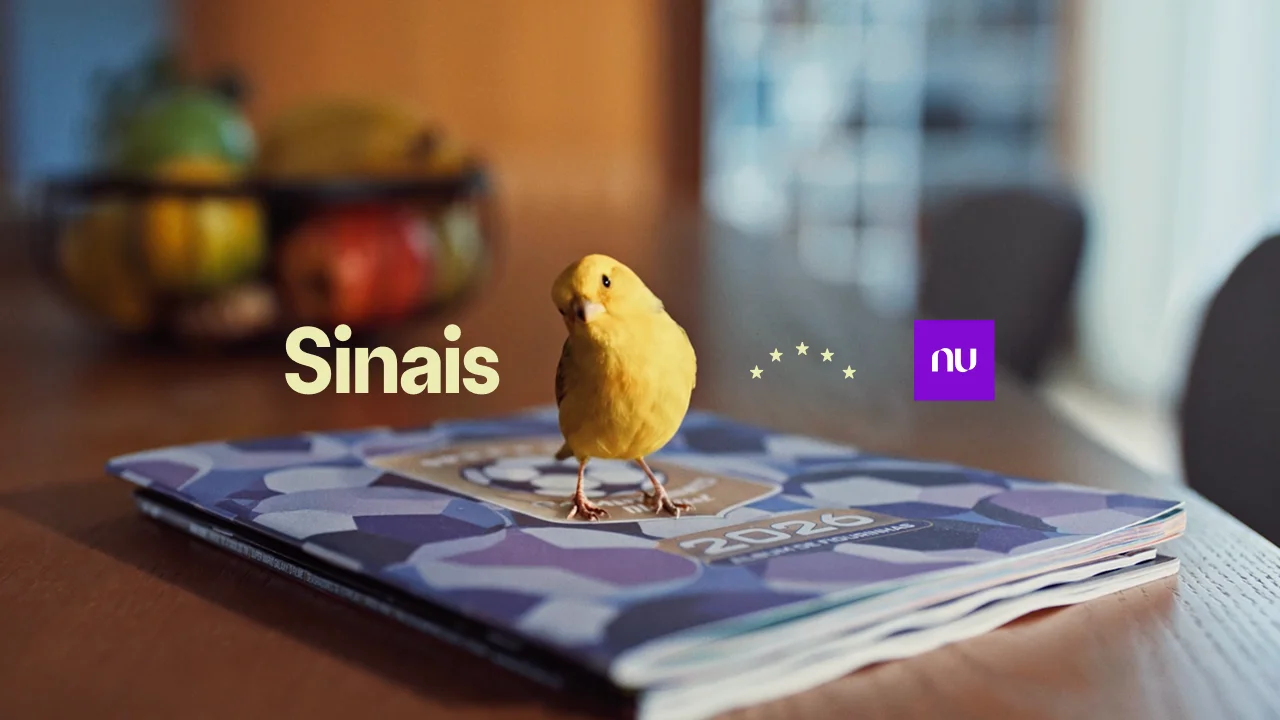 Um canário amarelo pousado sobre uma pilha de álbuns de figurinhas em uma mesa de madeira, com a palavra "Sinais" em letras grandes cor creme à esquerda, estrelas douradas ao centro e o logo roxo do Nubank à direita.