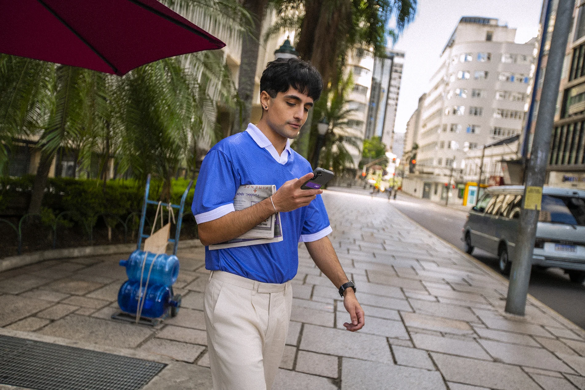 Homem jovem, branco, de cabelo escuro, caminha por uma calçada urbana usando camiseta azul e calça bege. Segura um celular com capinha roxa do Nubank e carrega um jornal sob o braço. Ao fundo, prédios, palmeiras e guarda-sol vinho.