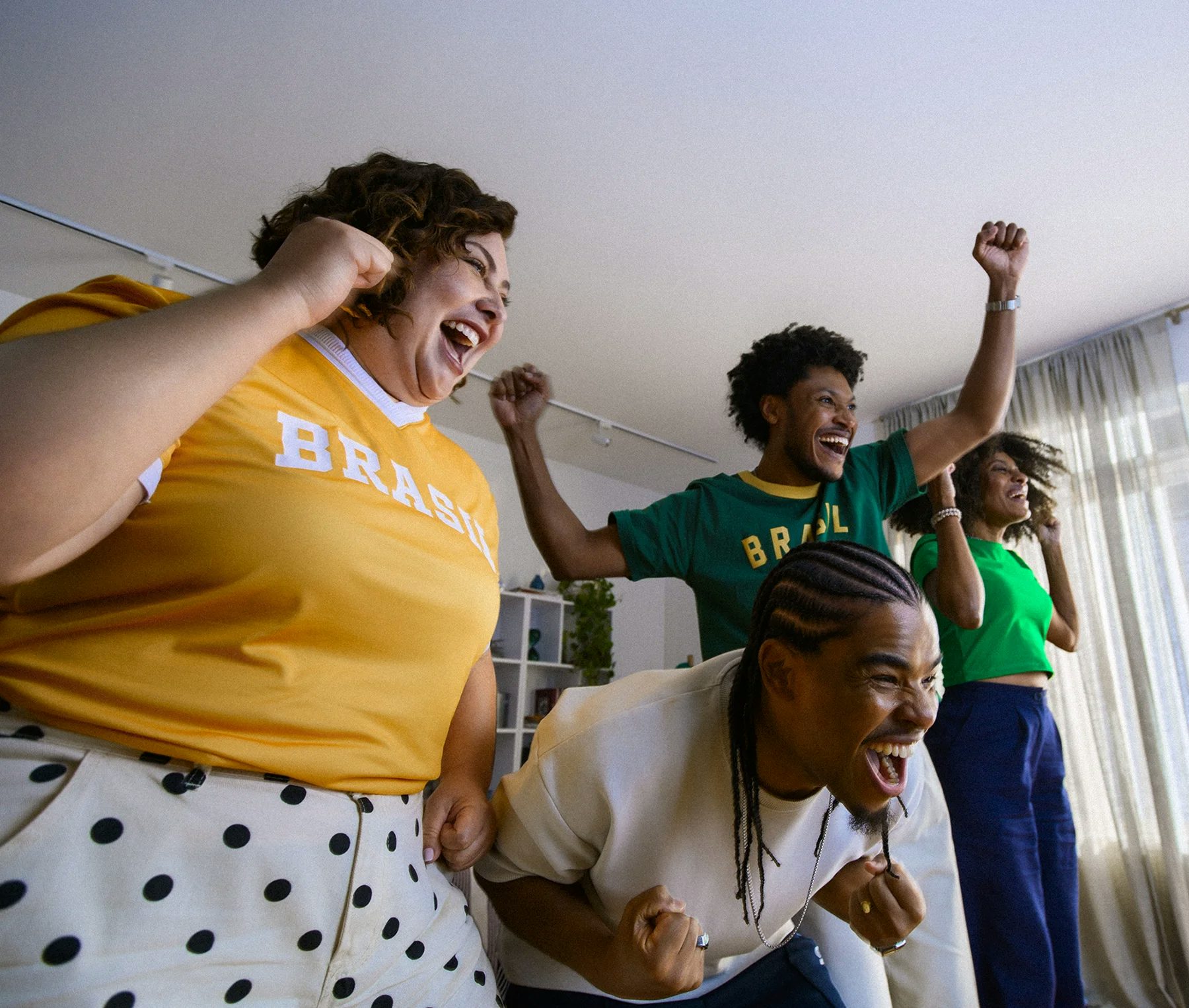 Quatro pessoas comemoram animadamente em uma sala de estar. À esquerda, uma mulher branca de cabelo cacheado usa camiseta amarela com "Brasil" e calça branca de poás. Ao centro, um homem negro com tranças se inclina para frente celebrando. Ao fundo, um homem negro com camiseta verde "Brasil" ergue o punho, e uma mulher negra de top verde sorri. Estante branca e cortinas ao fundo.