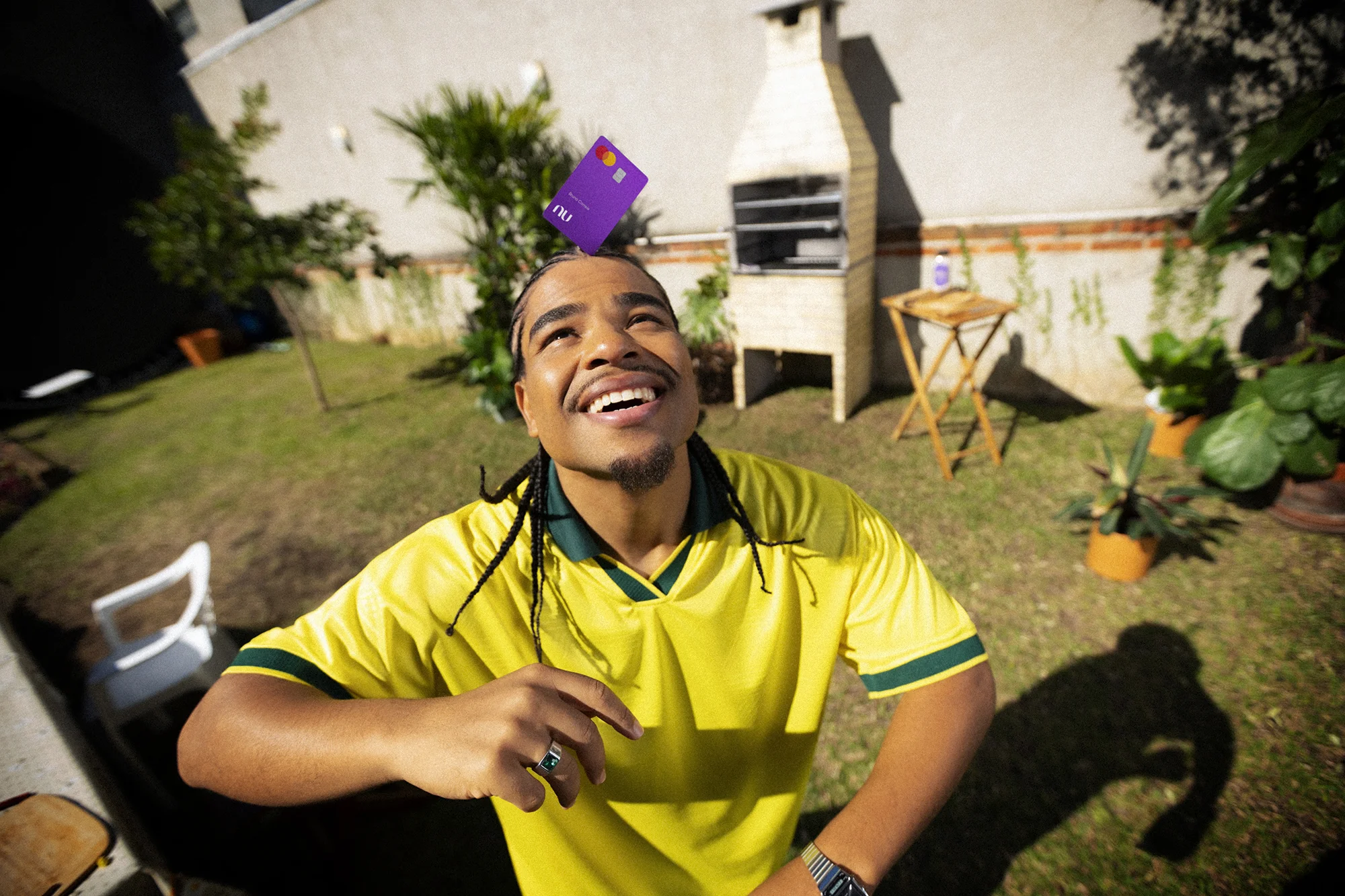 Homem jovem, negro, com tranças, usando camisa amarela da seleção brasileira, sorri e olha para cima enquanto equilibra um cartão roxo do Nubank sobre a cabeça. Ao fundo, um quintal com churrasqueira de alvenaria, plantas e grama verde.