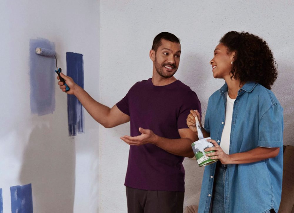Homem em pé, passa com sua mão direita um rolo de tinta azul em uma parede branca, enquanto olha para a esquerda e conversa com uma mulher negra segurando com a mão esquerda uma lata de tinta branca e com a direita um pincel.