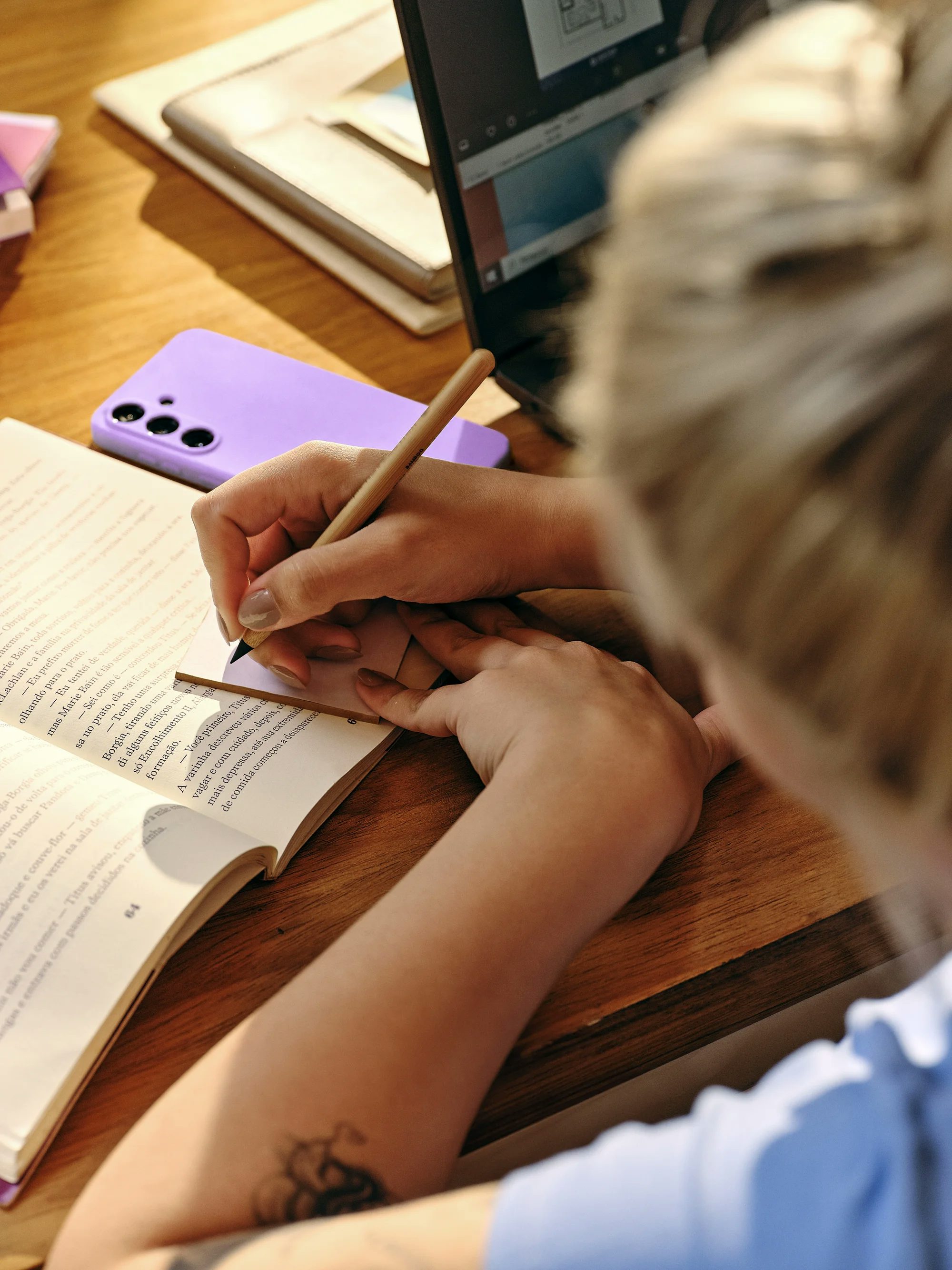 Uma pessoa aparece estudando em uma mesa de madeira, fazendo anotações com lápis em um pequeno bloco sobre um livro aberto. Ao redor, há um celular com capinha lilás e um notebook aberto, sob uma iluminação quente e acolhedora.
