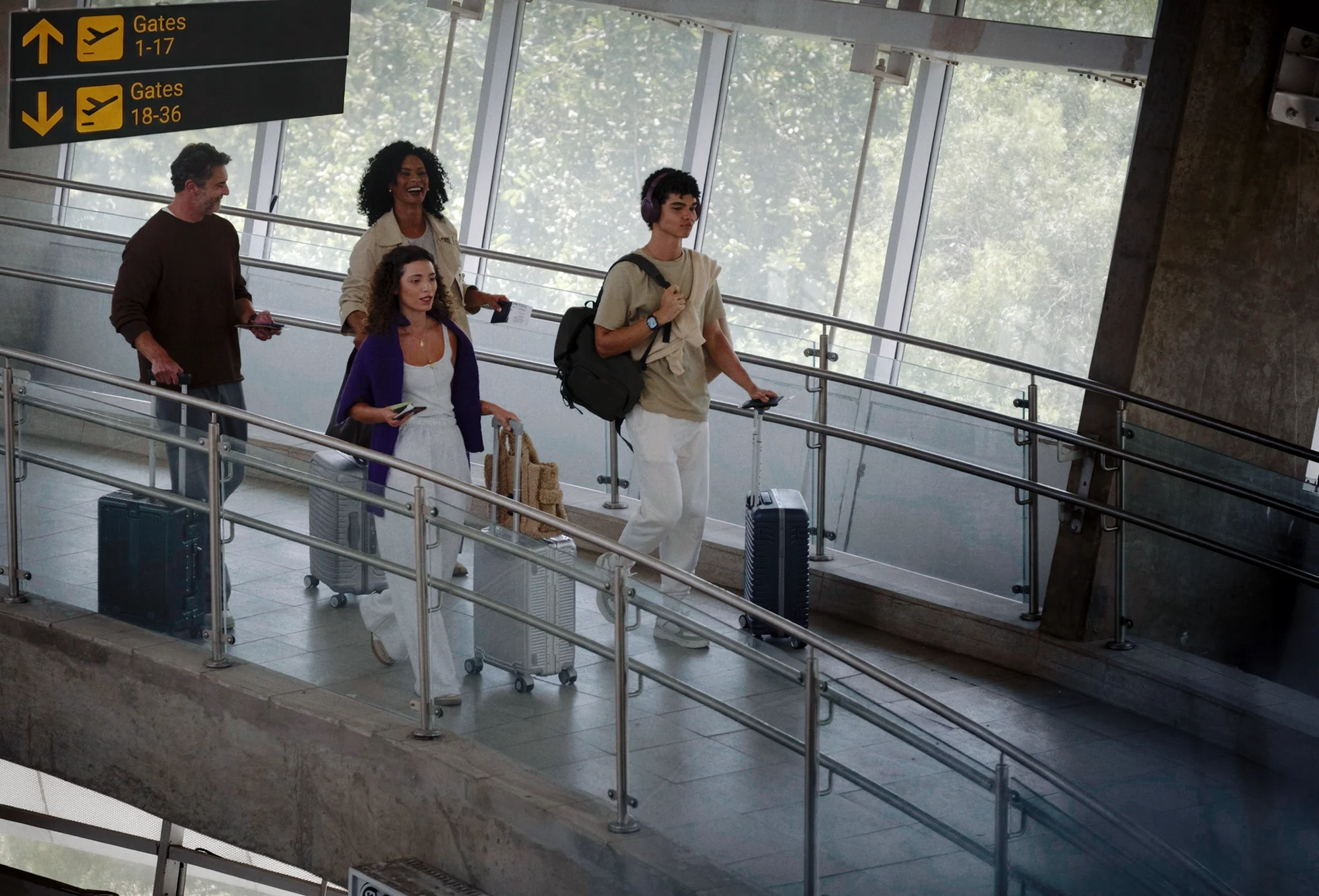 Um grupo de quatro pessoas caminha pelo corredor de um terminal de aeroporto. Eles puxam malas de rodinha prateadas e escuras, passando abaixo de uma placa de sinalização que indica os portões de embarque