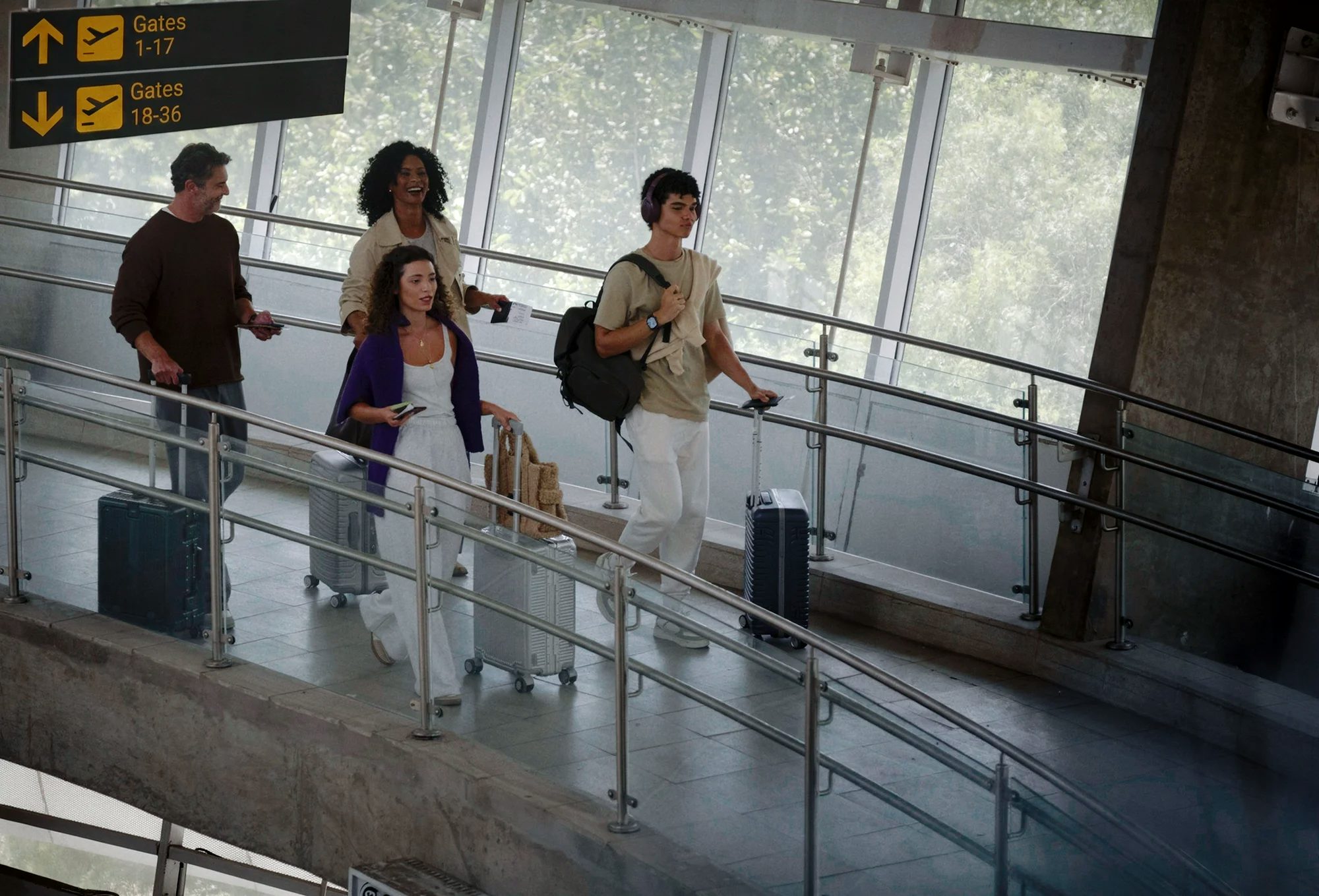 Um grupo de quatro pessoas caminha pelo corredor de um terminal de aeroporto. Eles puxam malas de rodinha prateadas e escuras, passando abaixo de uma placa de sinalização que indica os portões de embarque