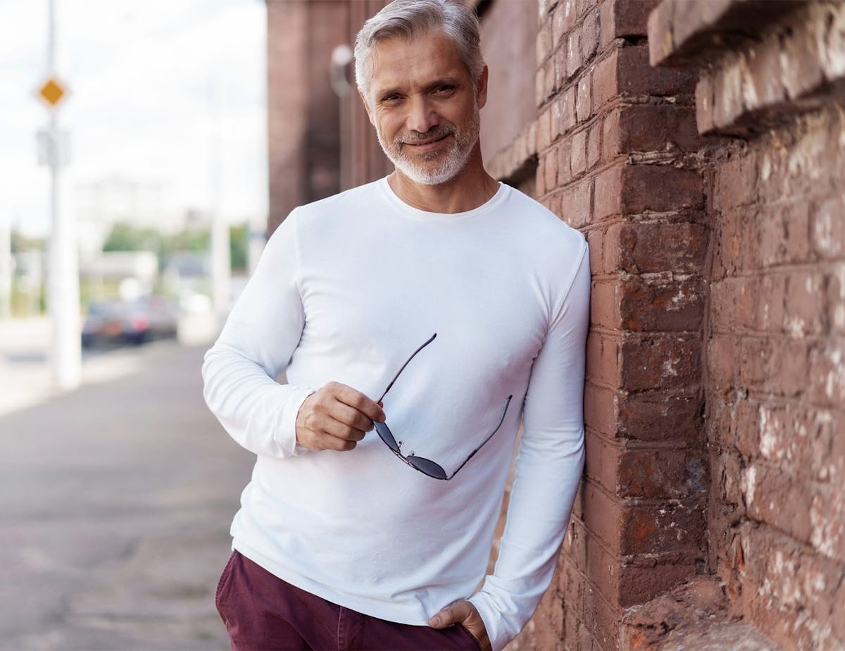 older man in white long sleeve leaning on brick wall