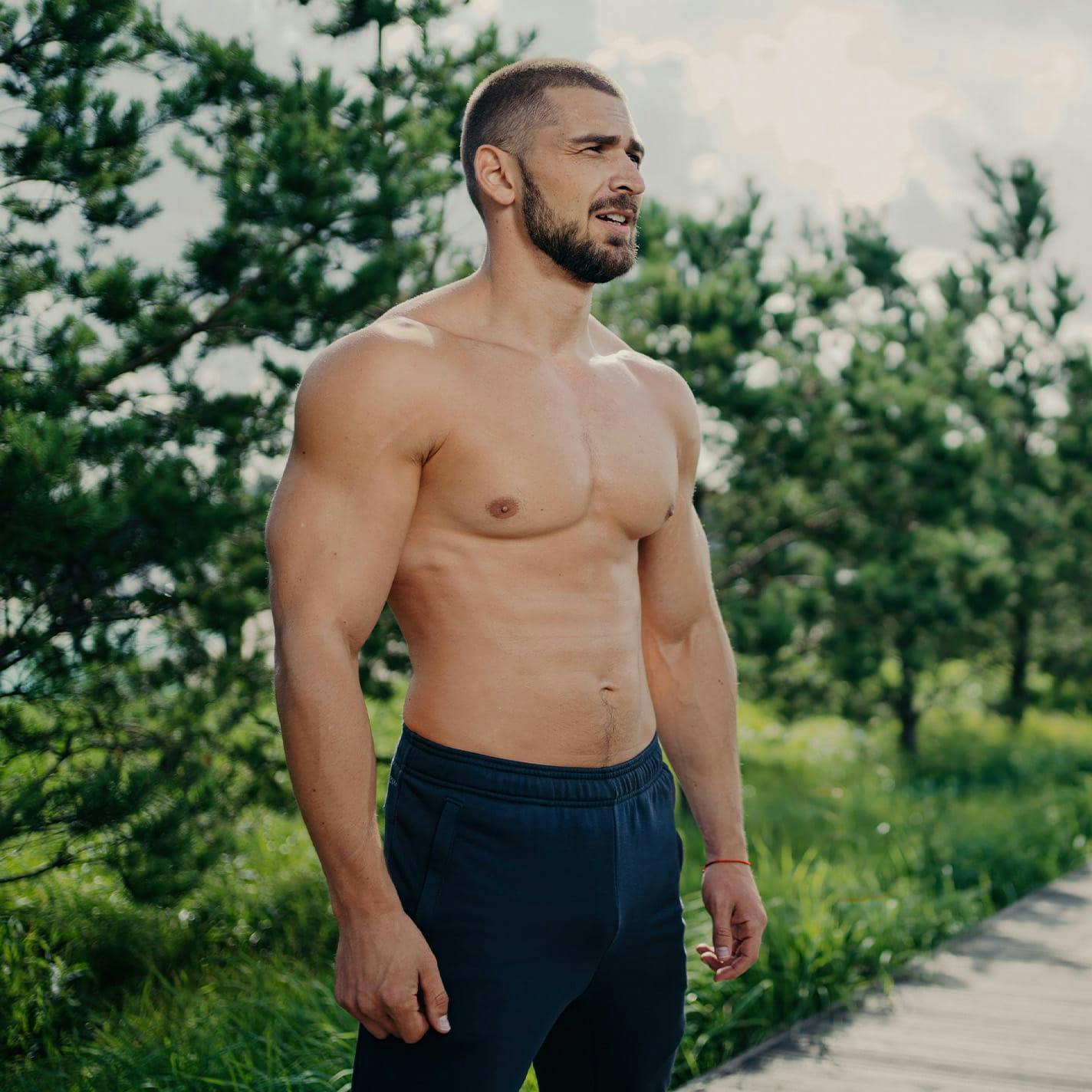 man with no shirt on standing in front of small trees