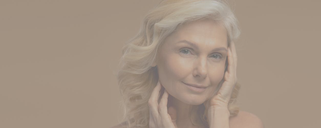 Older woman with blonde hair smiles while holding her hands to her face in a studio shoot. Her definited jaw line, and tight skin emulates the potential results of a Nashville Facelift with Dr. Higdon