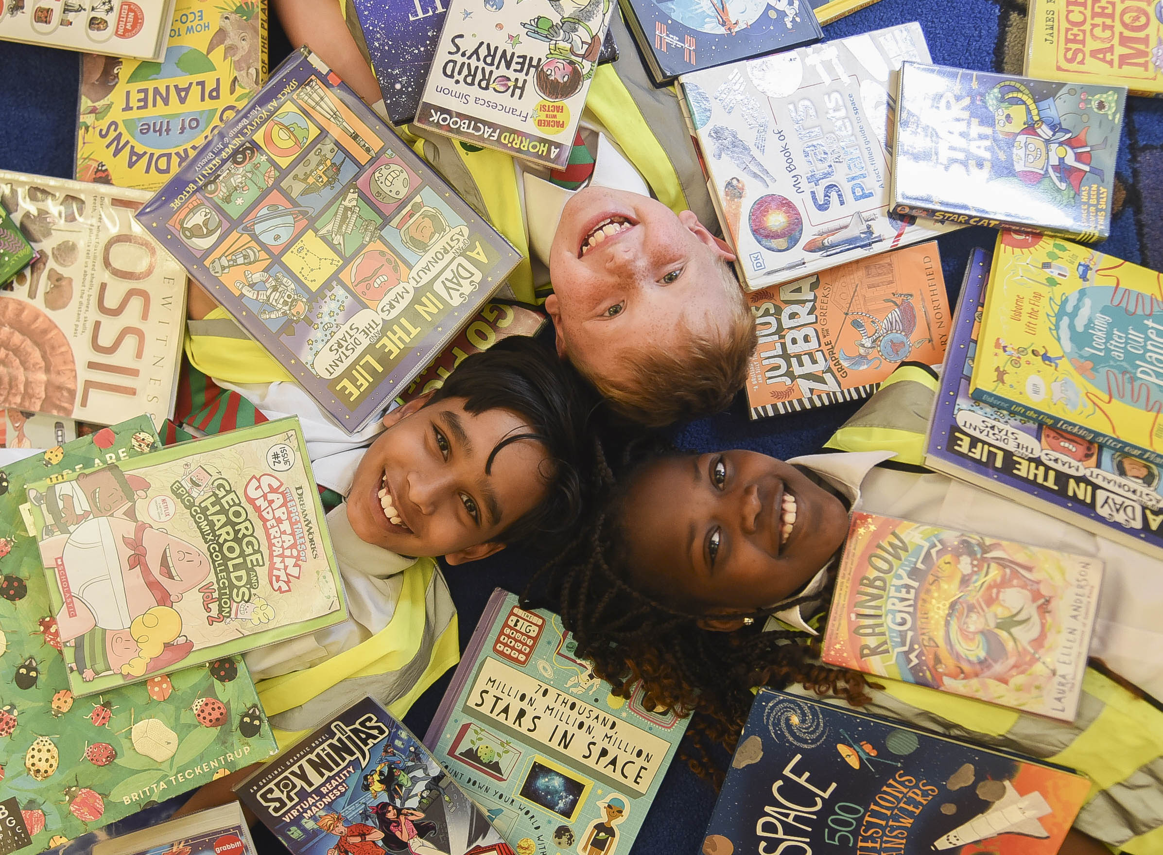 Three children lying on the floor of a library covered with books
