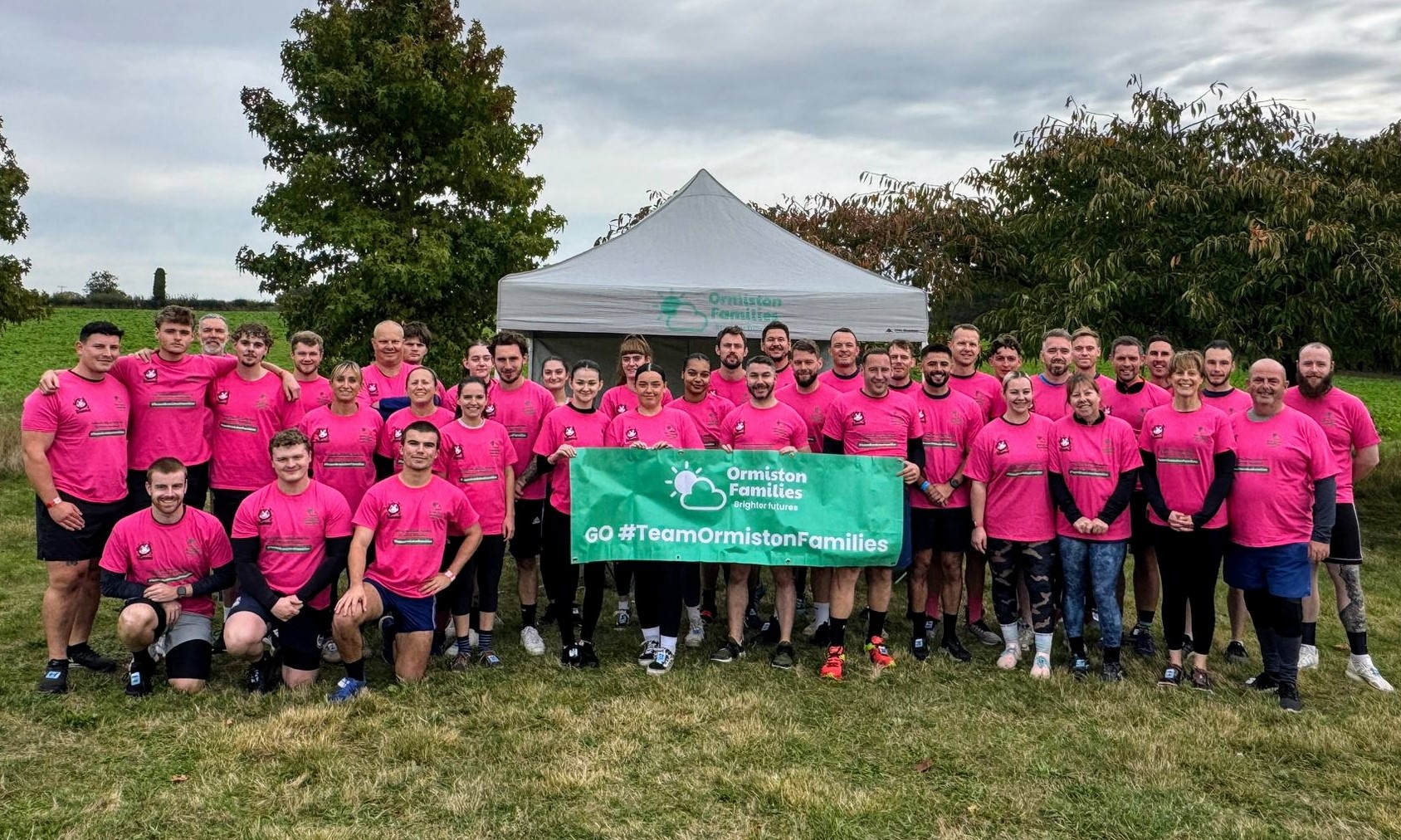 Large group of Ormiston Families volunteers wearing pink shirts standing together outdoors, holding a green banner reading “Go #TeamOrmistonFamilies” in front of a charity gazebo