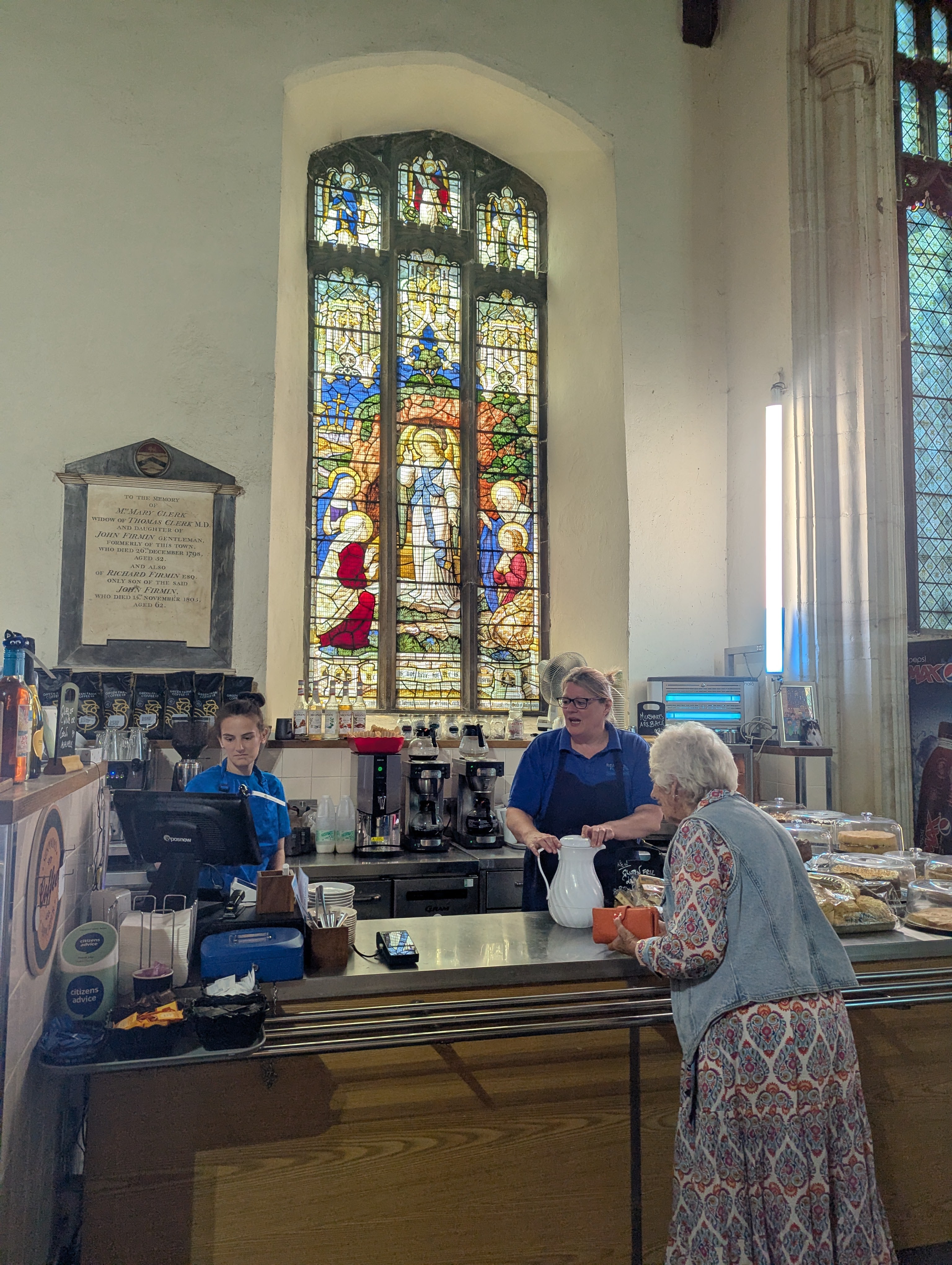 Two café staff serving an older customer at the counter in St Lawrence Café, with stained-glass windows behind them