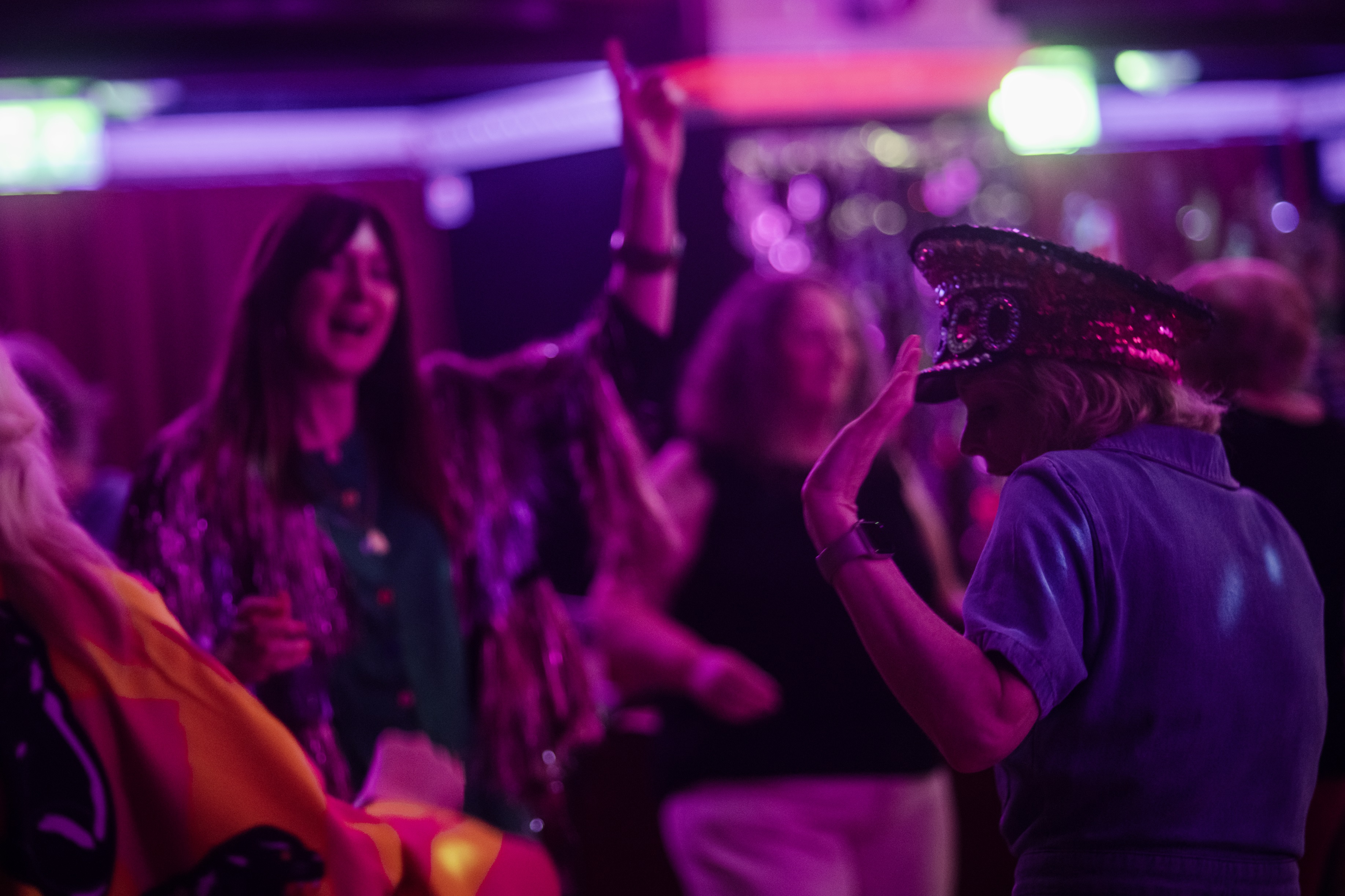 People dance under pink and purple lights at a Grief Disco, one woman wearing a sequinned disco hat