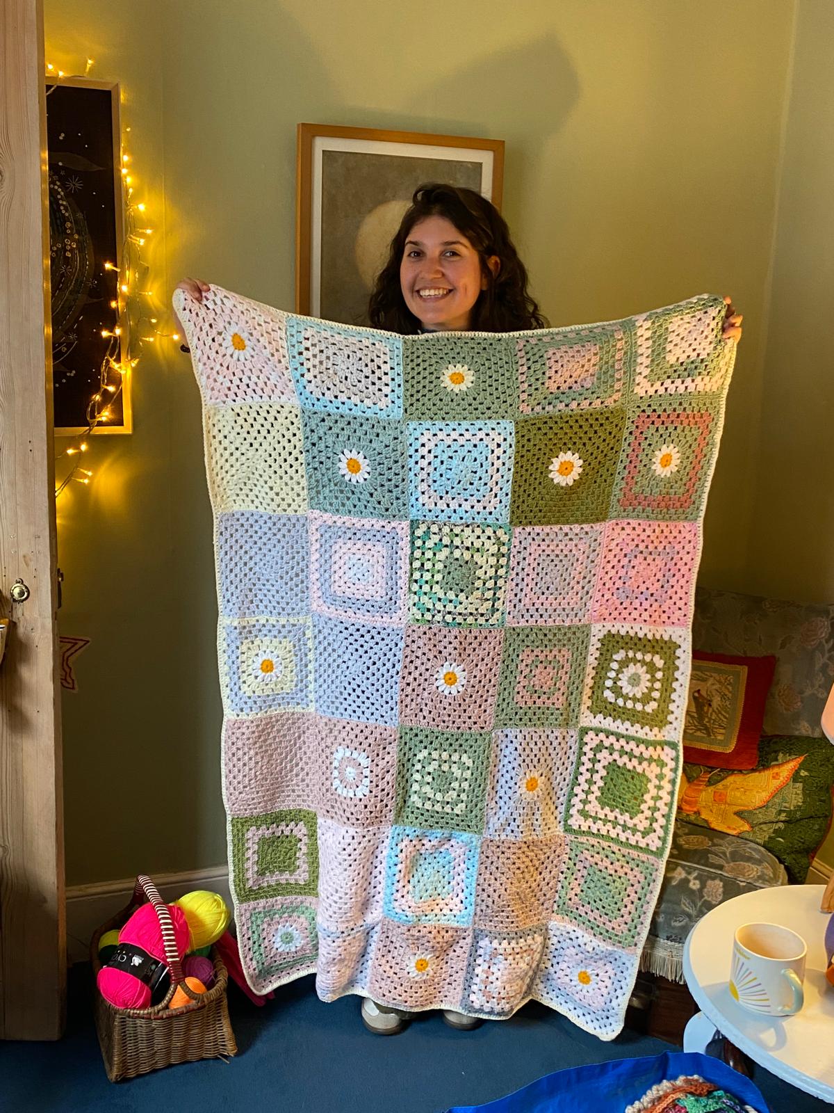 A woman smiling while holding up a completed crocheted blanket of pastel-coloured squares with daisy patterns