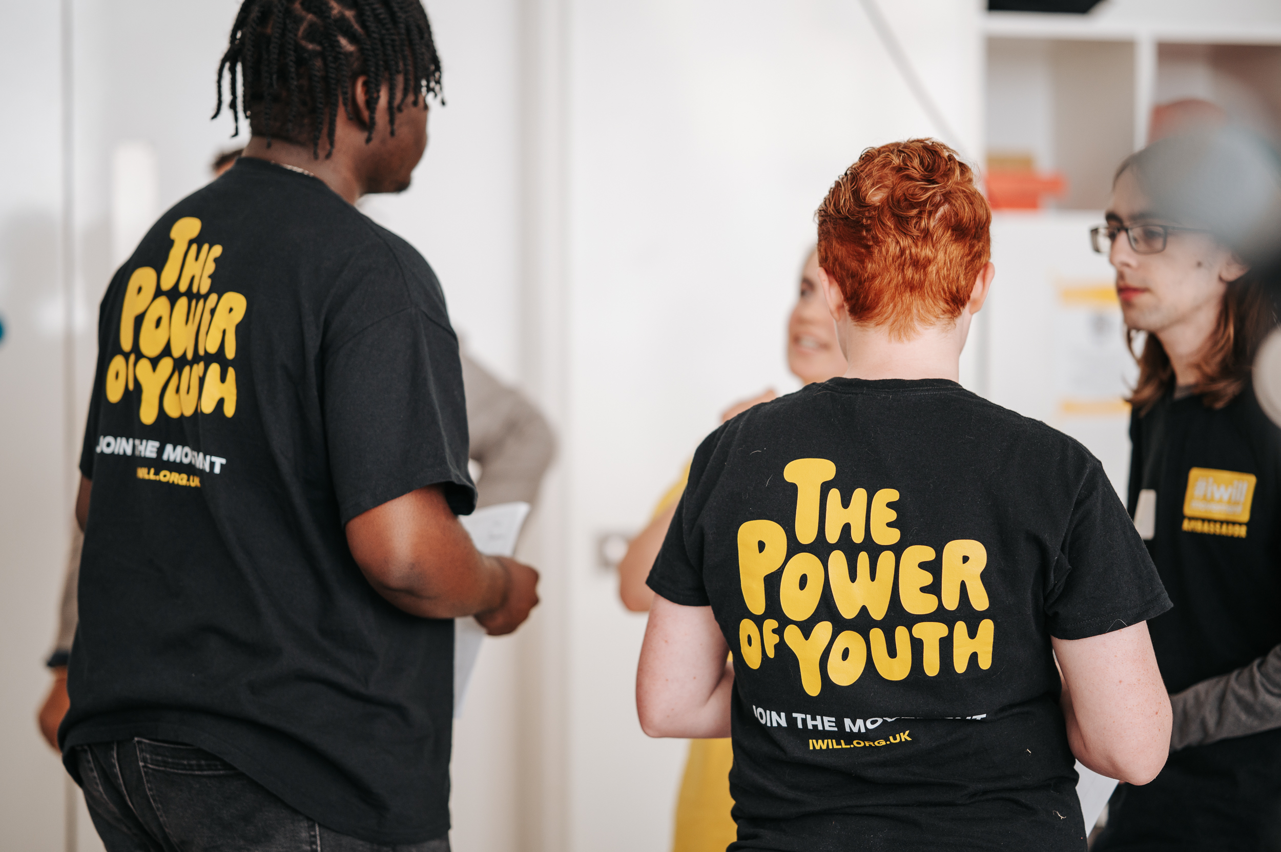 Back view of young people wearing black t-shirts with yellow text reading "THE POWER OF YOUTH" and "JOIN THE MOVEMENT #IWILL.ORG.UK"
