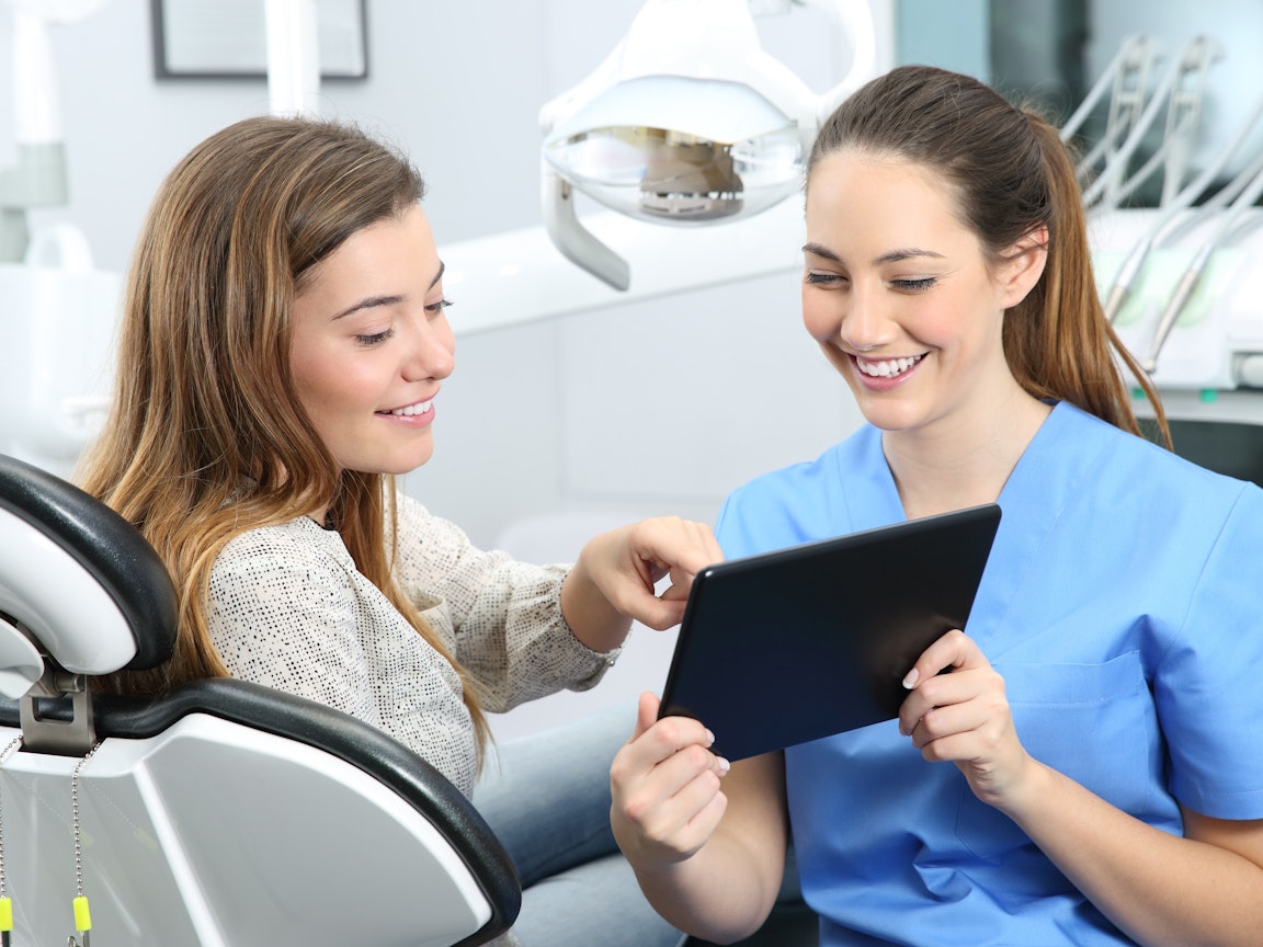 Junge Frau auf Zahnarztstuhl bespricht etwas auf dem Tablet mit der Ärztin