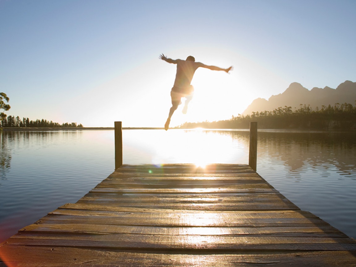 Mann springt bei Sonnenaufgang vom Steg in einen See Mann springt bei Sonnenaufgang vom Steg in einen See