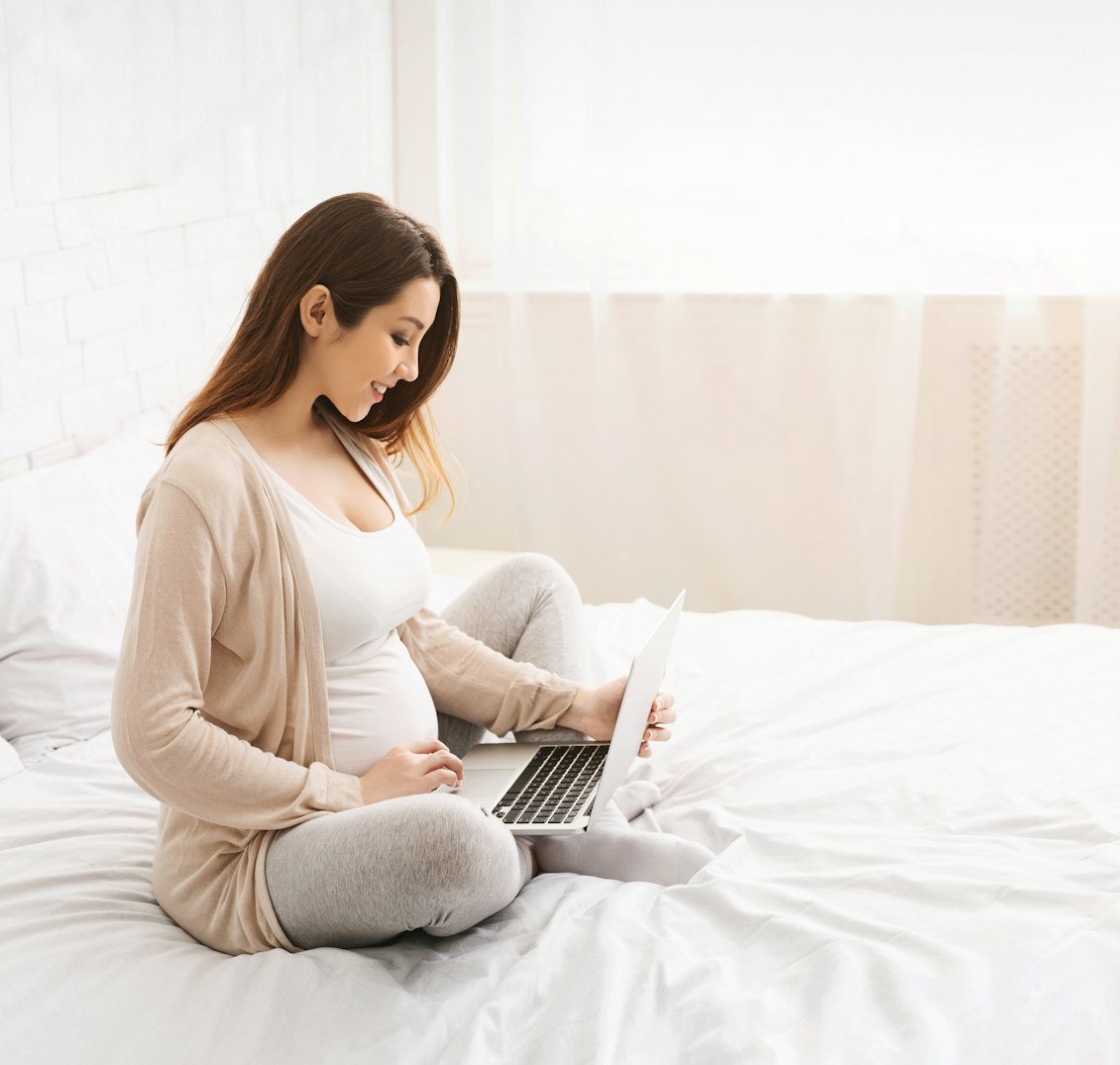 Schwangere Frau sitzt mit einem Laptop auf dem Bett