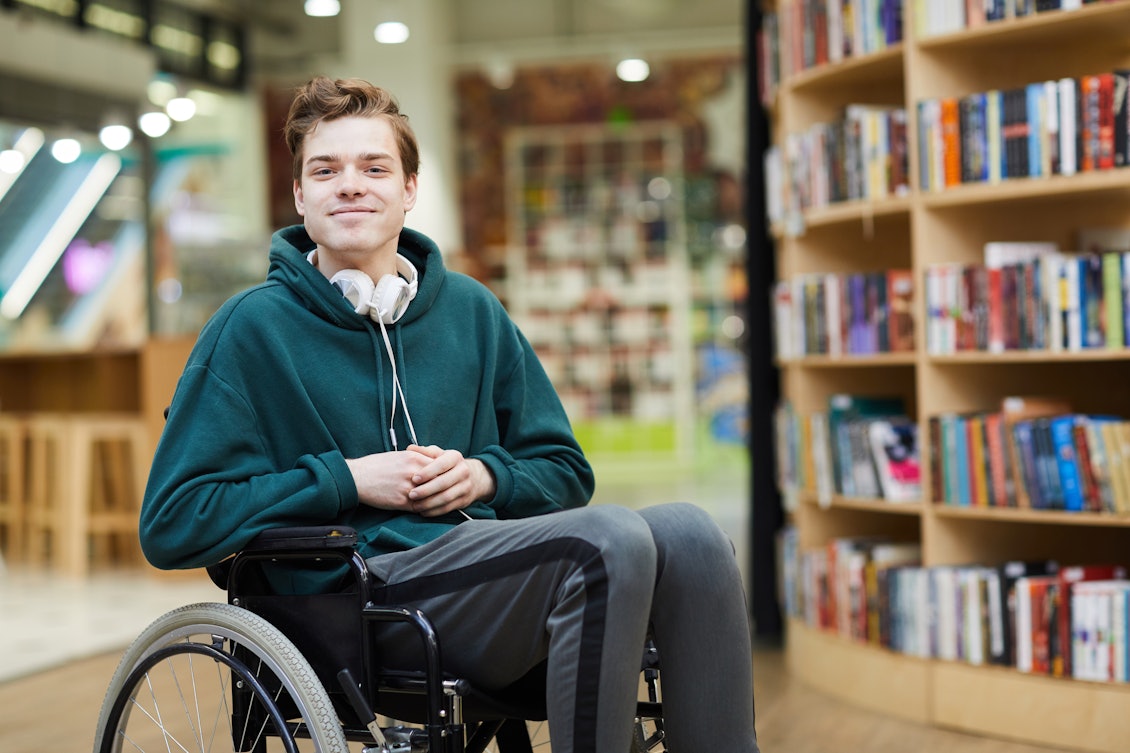 Junger gutaussehender Mann mit grünem Pullover sitzt in seinem Rollstuhl in einer Bibliothek
