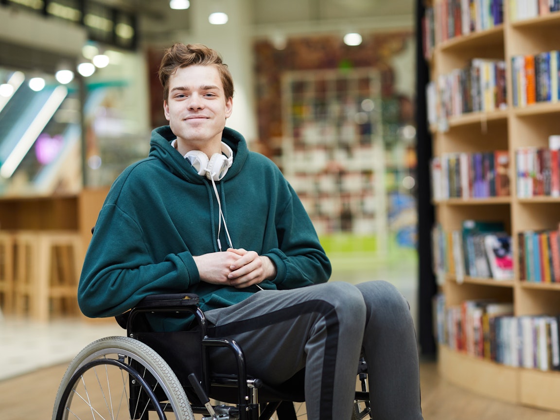 Junger Mann im Rollstuhl Junger gutaussehender Mann mit grünem Pullover sitzt in seinem Rollstuhl in einer Bibliothek
