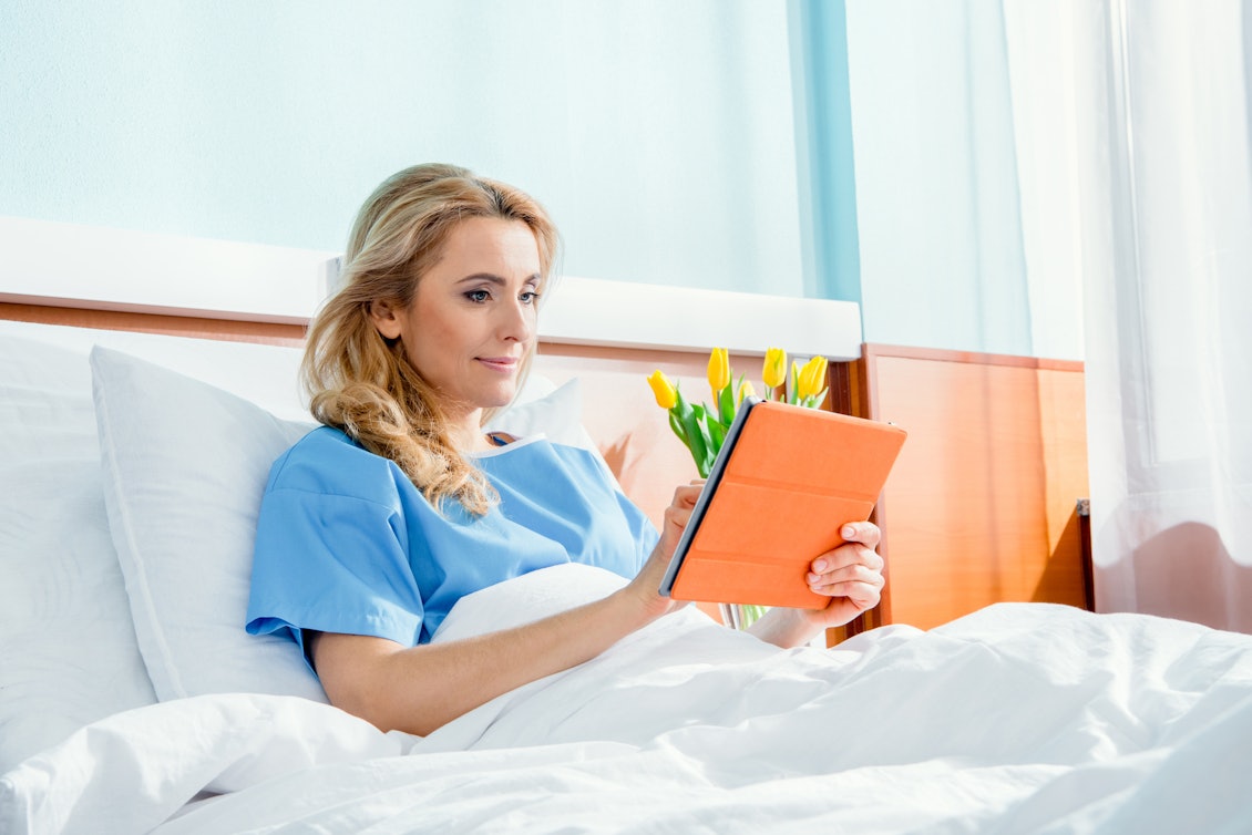 Junge blonde Frau sitzt im Krankenbett und arbeitet am Tablet