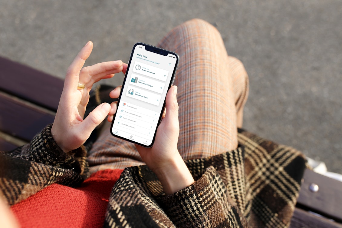 Frau hat Handy mit TeleClinic App in der Hand und sitzt auf einer Parkbank Frau hat Handy mit TeleClinic App in der Hand und sitzt auf einer Parkbank