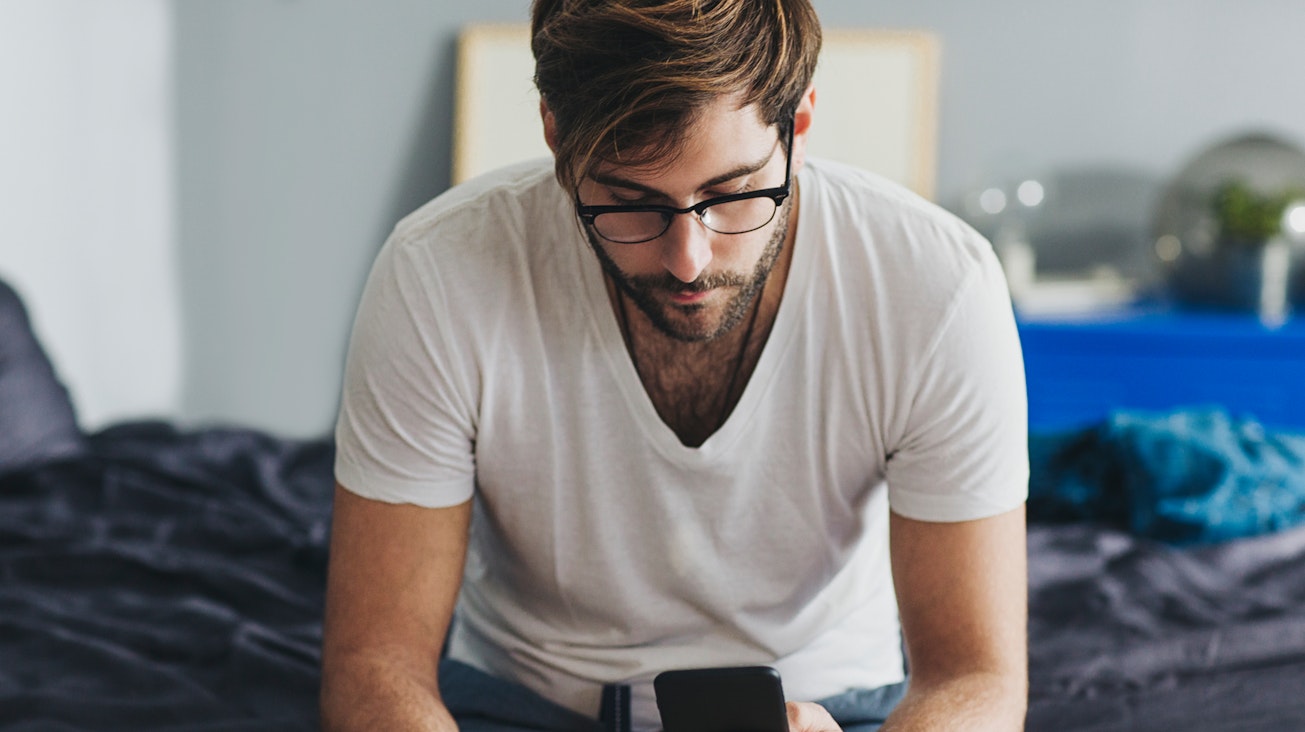 Mann mit Handy auf Bett Ein junger Mann nutzt sein Handy und sitzt auf dem Bett