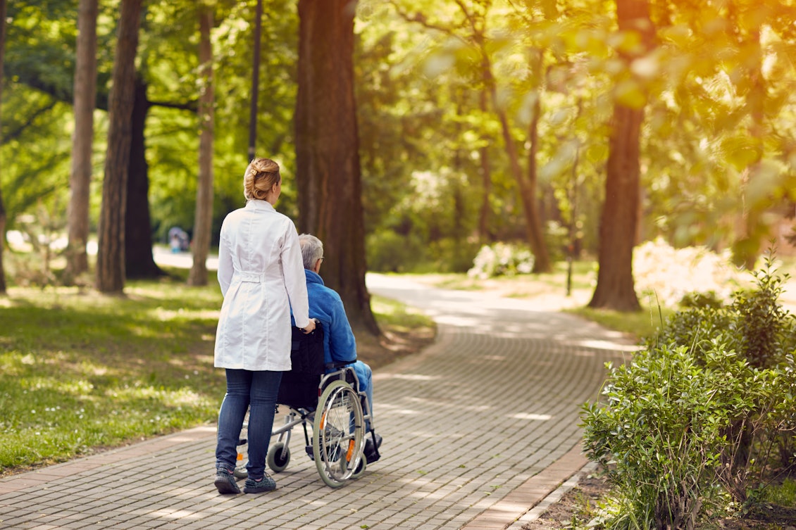 Eine Pflegerin schiebt einen Mann im Rollstuhl durch einen schönen Park