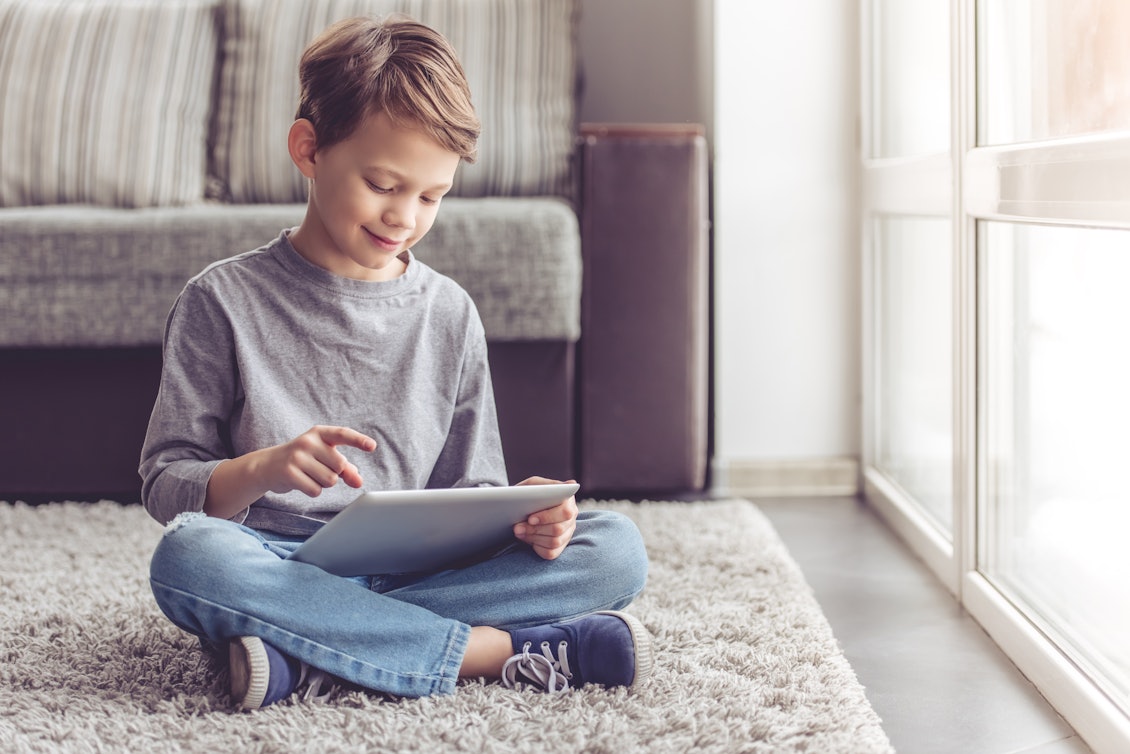 Eine Zusatzversicherung lohnt sich bereits in jungen Jahren Ein Junge sitzt im Schneidersitz auf dem Boden und spielt mit einem Tablet.