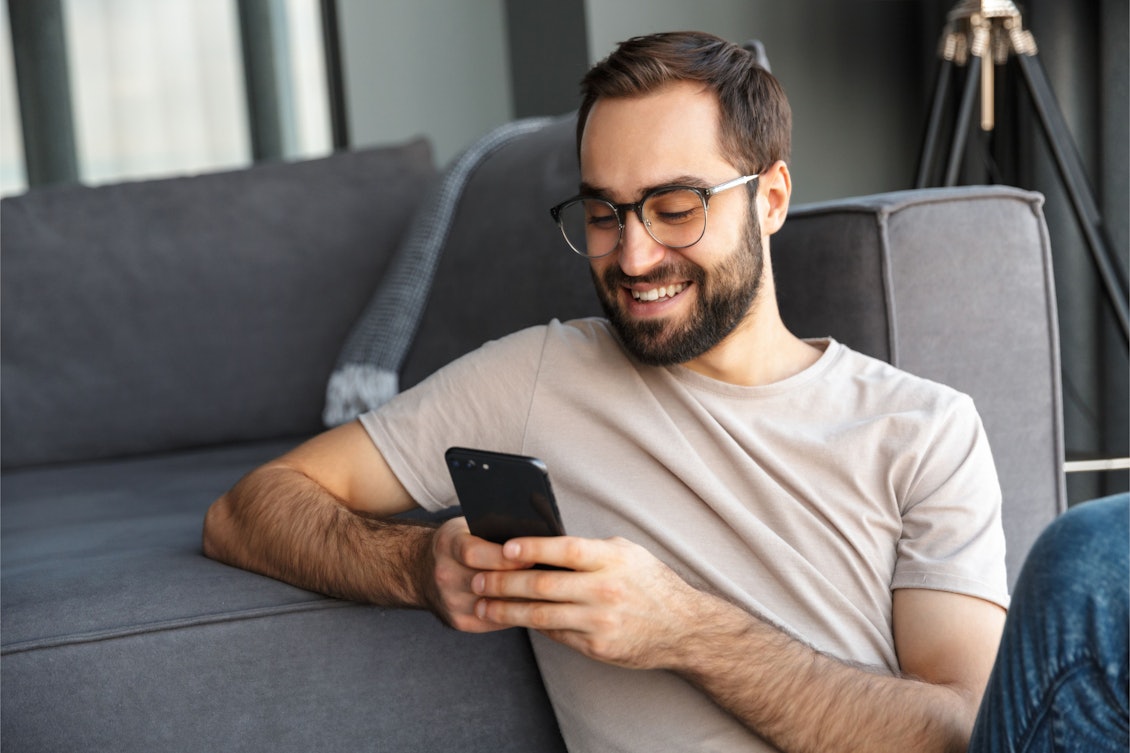 Junger Mann mit Handy sitzt auf dem Boden vor der Couch Mann mit Handy sitzt vor dem Sofa