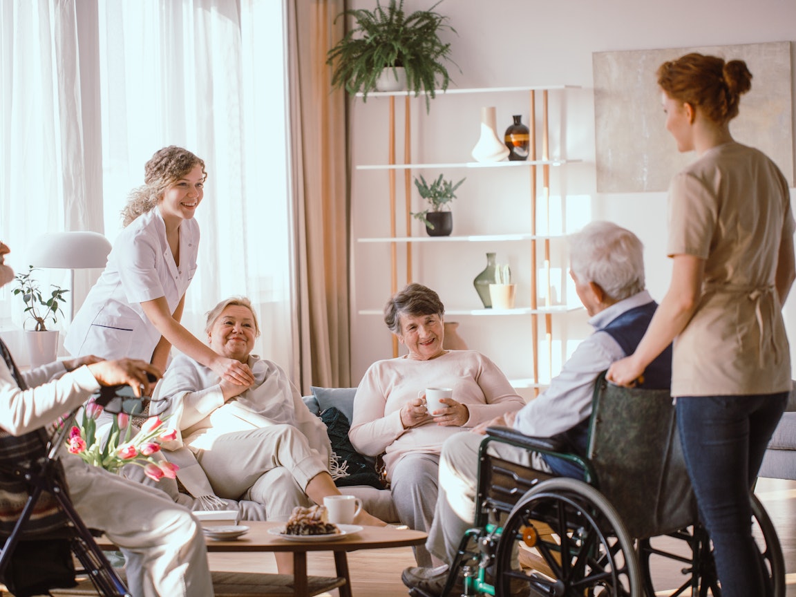Pflegebedürftige Personen haben Anspruch auf den Wohngruppenzuschlag Eine Gruppe Senioren und zwei Pflegekräfte sitzen gemütlich beisammen