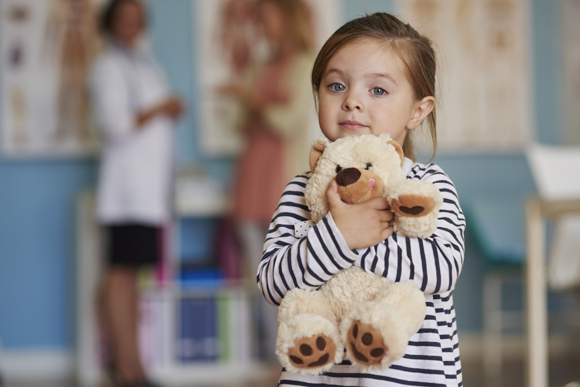 Ein kleines Mädchen ist mit seiner Mutter beim Arzt und hält einen Teddybär in der Hand.