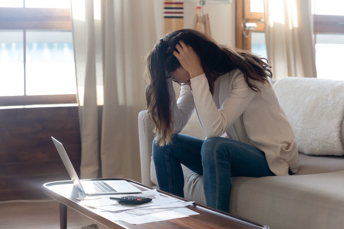 Junge Frau sitzt verzweifelt auf Sofa Junge Frau sitzt mit gesenktem Kopf auf dem Sofa vor ihrem Laptop