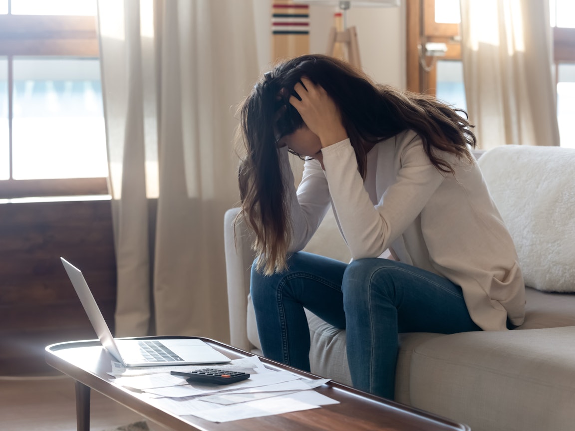 Junge Frau sitzt verzweifelt auf Sofa Junge Frau sitzt mit gesenktem Kopf auf dem Sofa vor ihrem Laptop