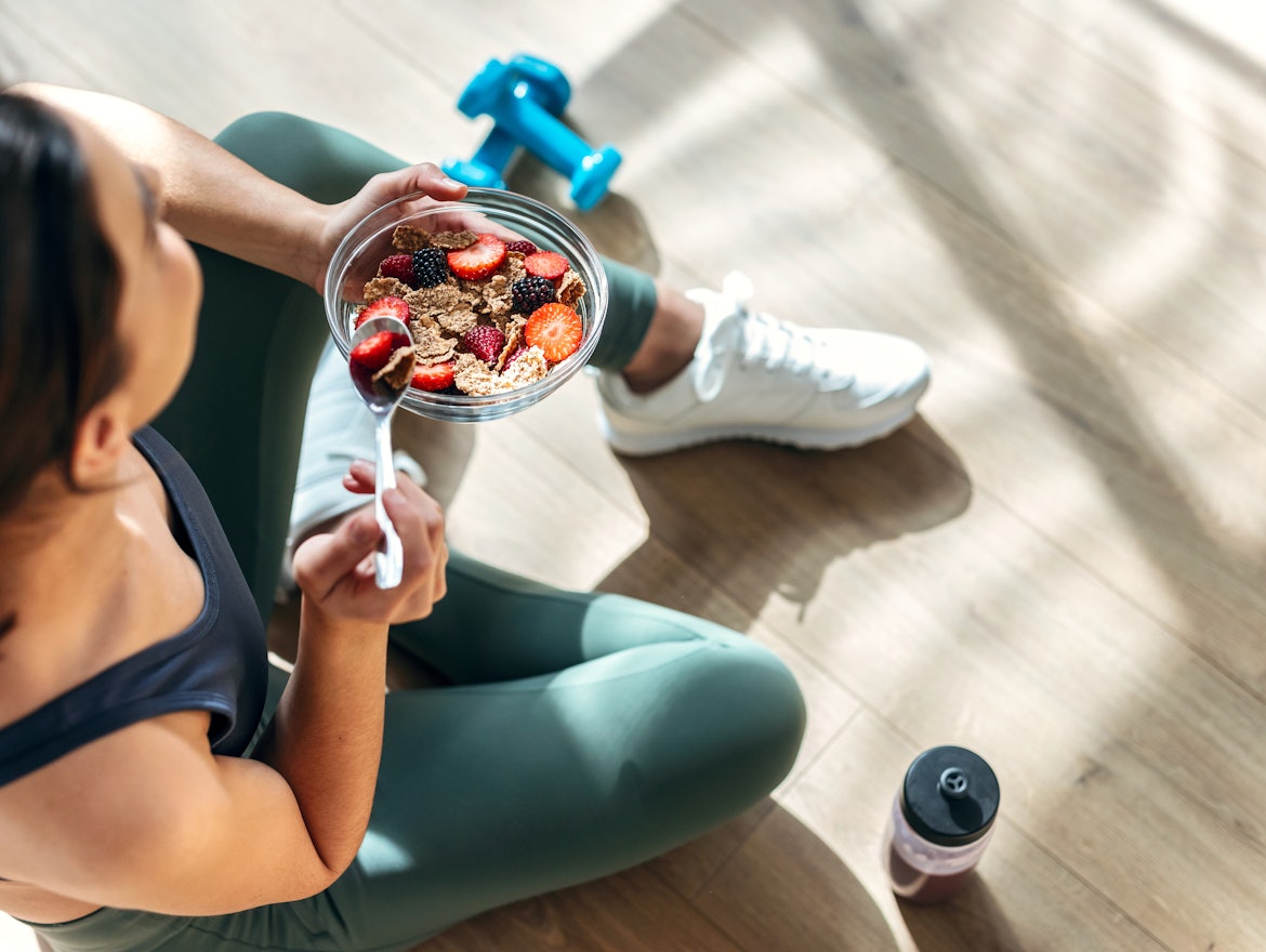 Frau sitzt mit Sportkleidung auf dem Boden und isst gesundes Müsli