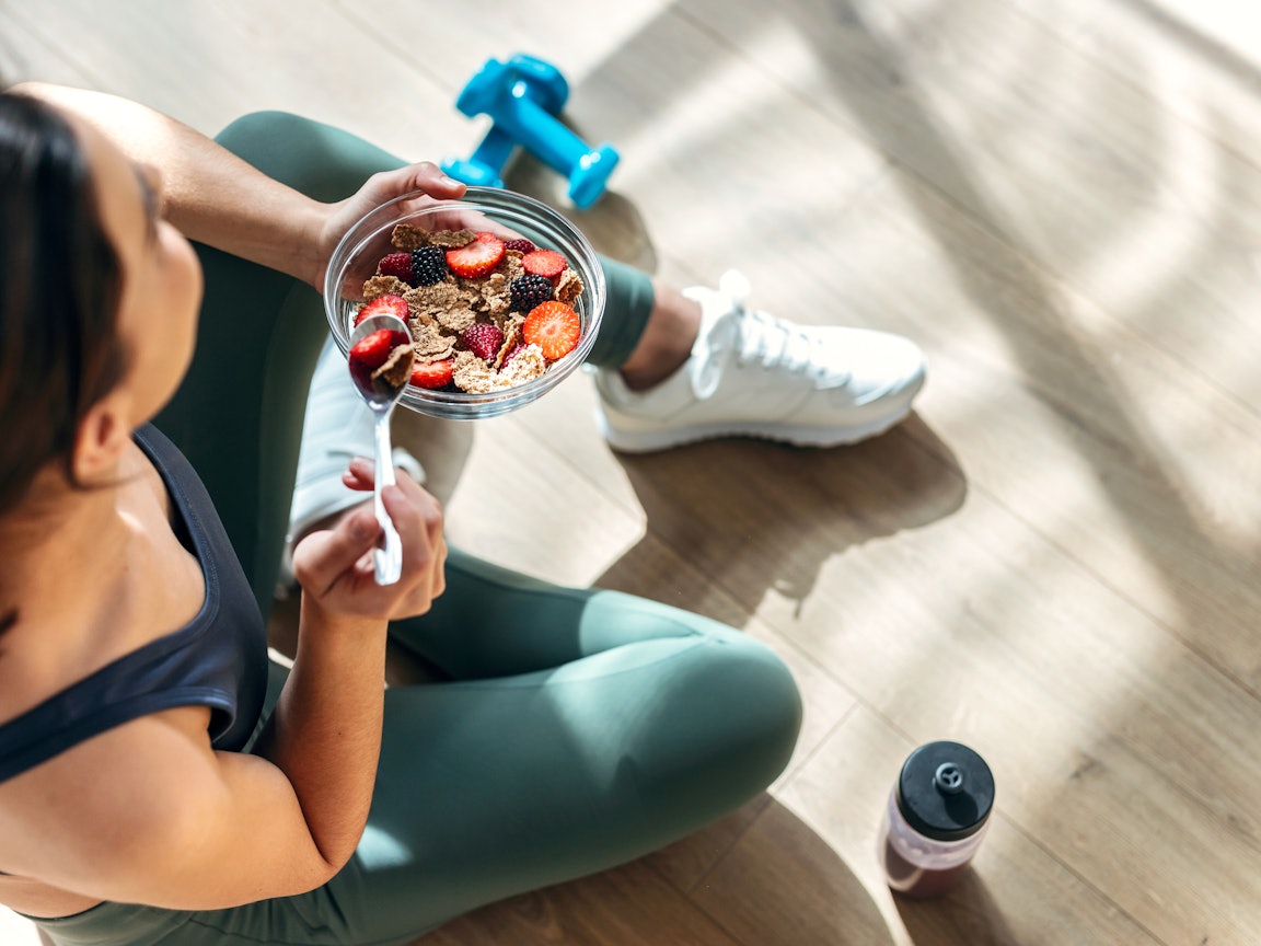 Frau in Sportkleidung mit gesundem Müsli Frau sitzt mit Sportkleidung auf dem Boden und isst gesundes Müsli