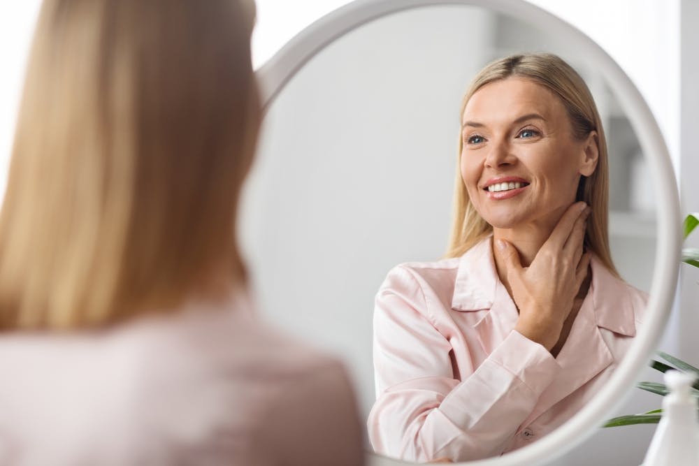 A smiling woman examines her neck, reflecting the benefits of local anesthesia in neck lift surgery.