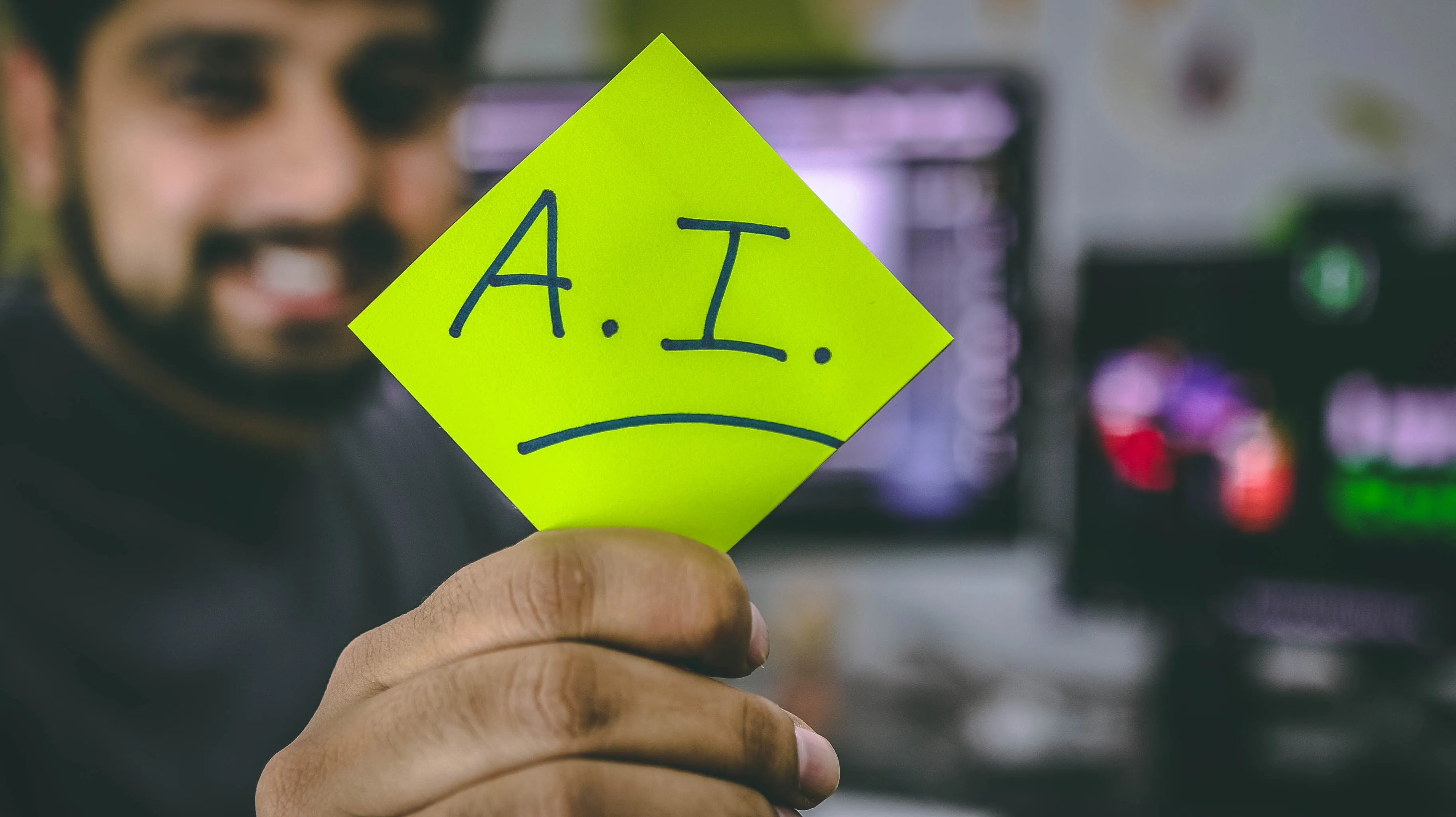 A photo of a man holding up a sticky-note that reads "AI"