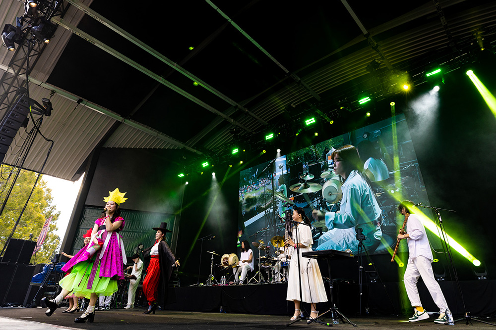 A band of stage with coloured lights and a large screen with a projection of the performance behind them