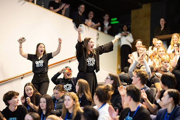 Hannah Duder standing up in an auditorium of people with her arms raised as she celebrates her team's win
