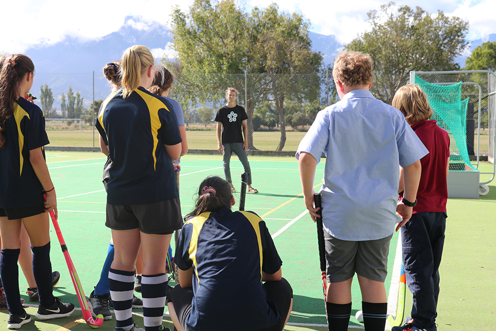 A group of kids in their hockey gear standing on a turf listening to Richard
