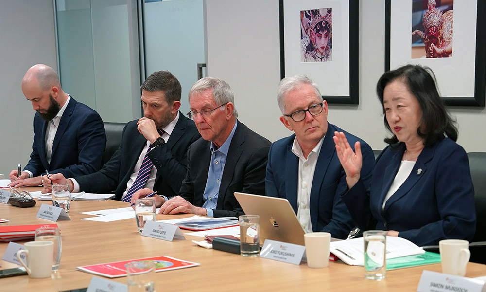 A woman speaking at a boardroom table with four people in the shot listening