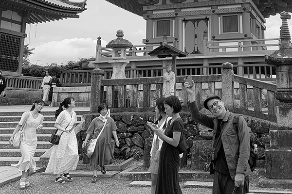 Daniel Belton waving to the camera in front of a shrine with women wealking by
