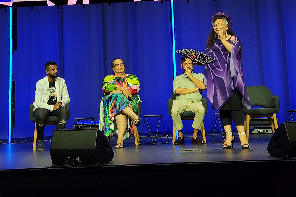 A woman on stage speaking into a microphone with three people sitting on chairs behind her