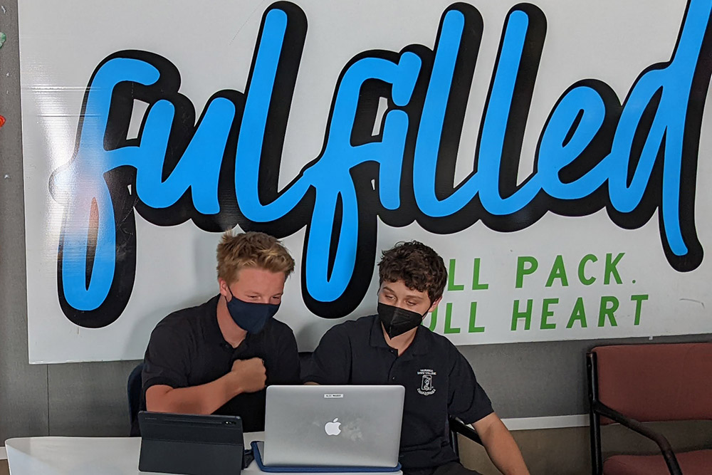 Two boys looking at a laptop with a banner saying 'Fulfilled behind them